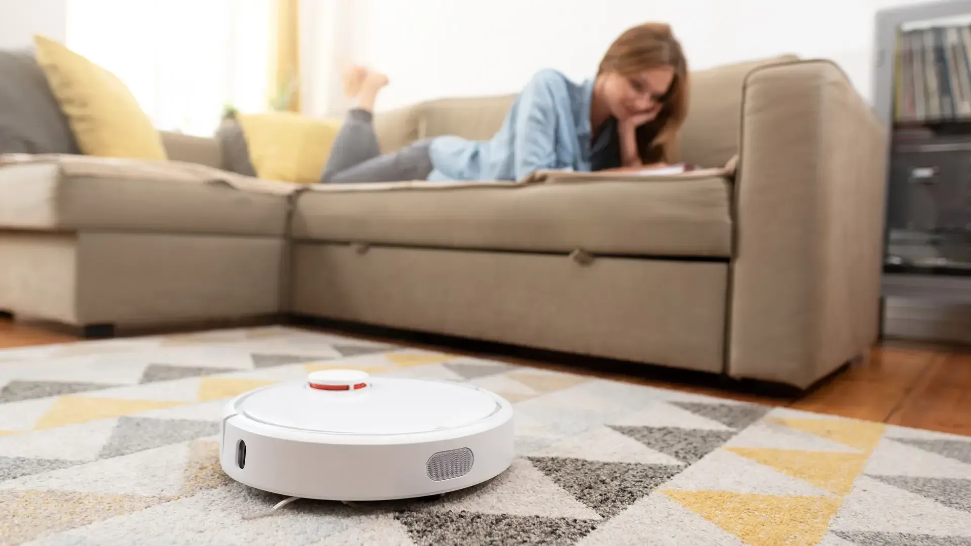 A woman is laying on a couch next to a robot vacuum cleaner.