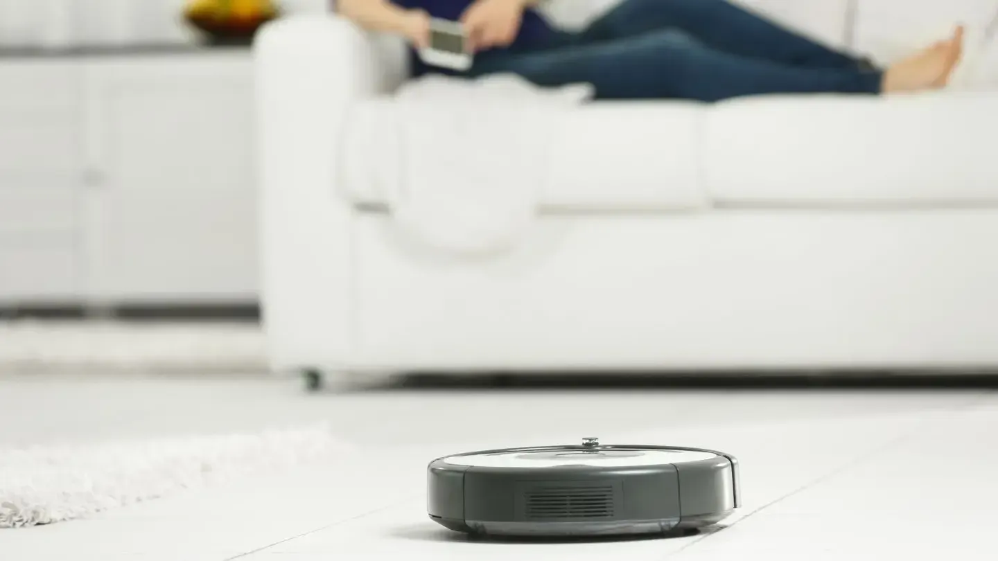 A person is sitting on a couch while a robot vacuum cleaner is cleaning the floor.