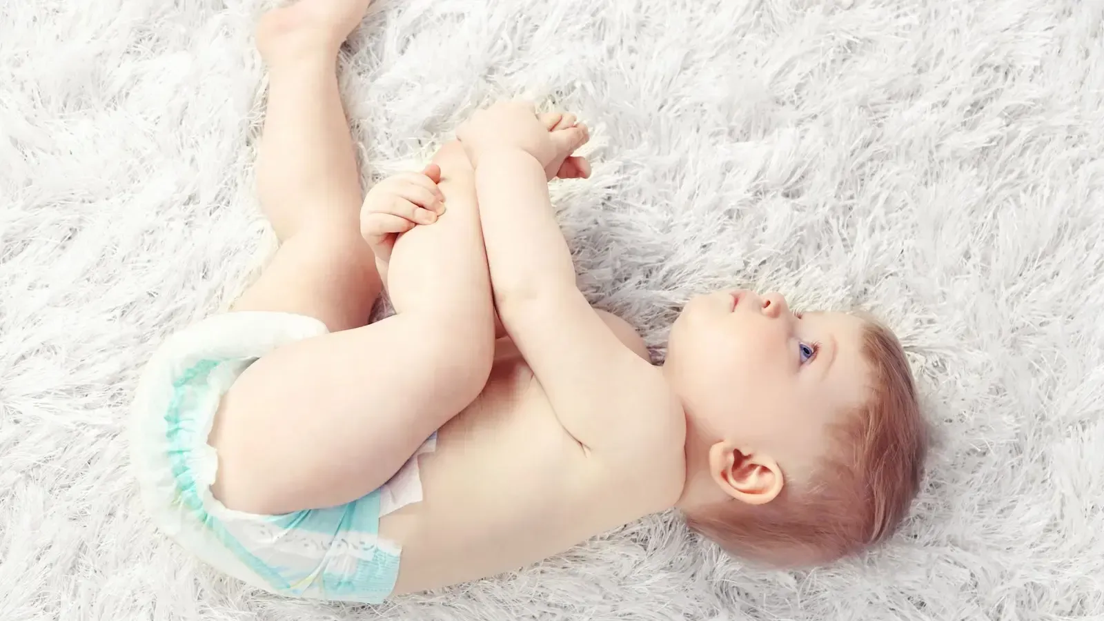 A baby in a diaper is laying on its back on a white carpet.