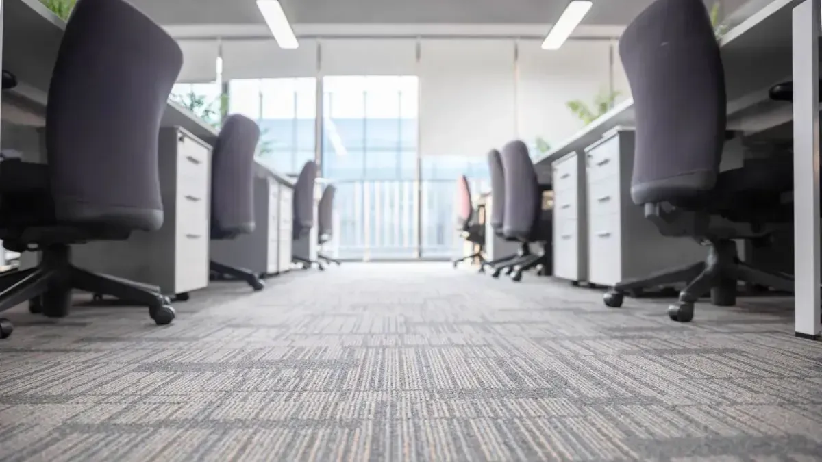 Office cubicles with grey carpet, chairs, and desks in front of a large window.