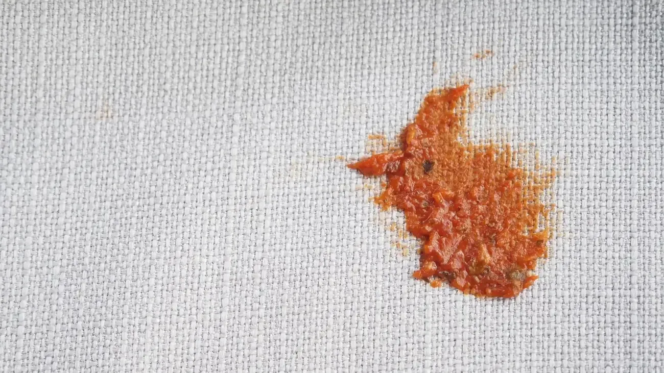 A textured light gray fabric surface with a fresh, reddish-brown sauce stain on the right side.