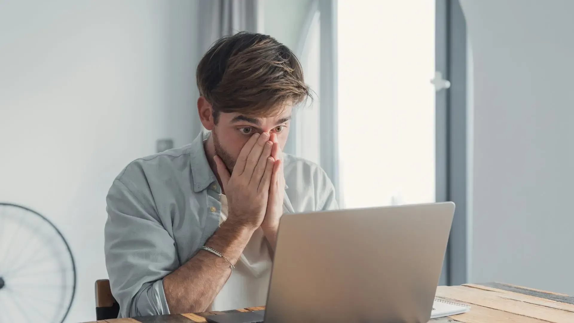 Man sitting at a laptop with hands over his face, looking stressed in a bright room
