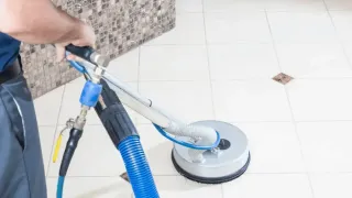 Person cleaning white tile floor with a circular cleaning machine and hoses.