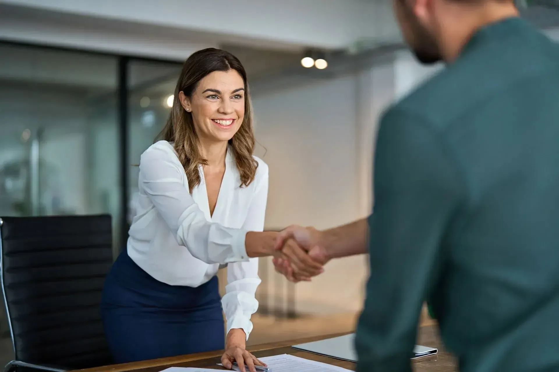 Woman in white shirt and blue skirt shaking hands with a person in a green shirt, smiling, in an office.