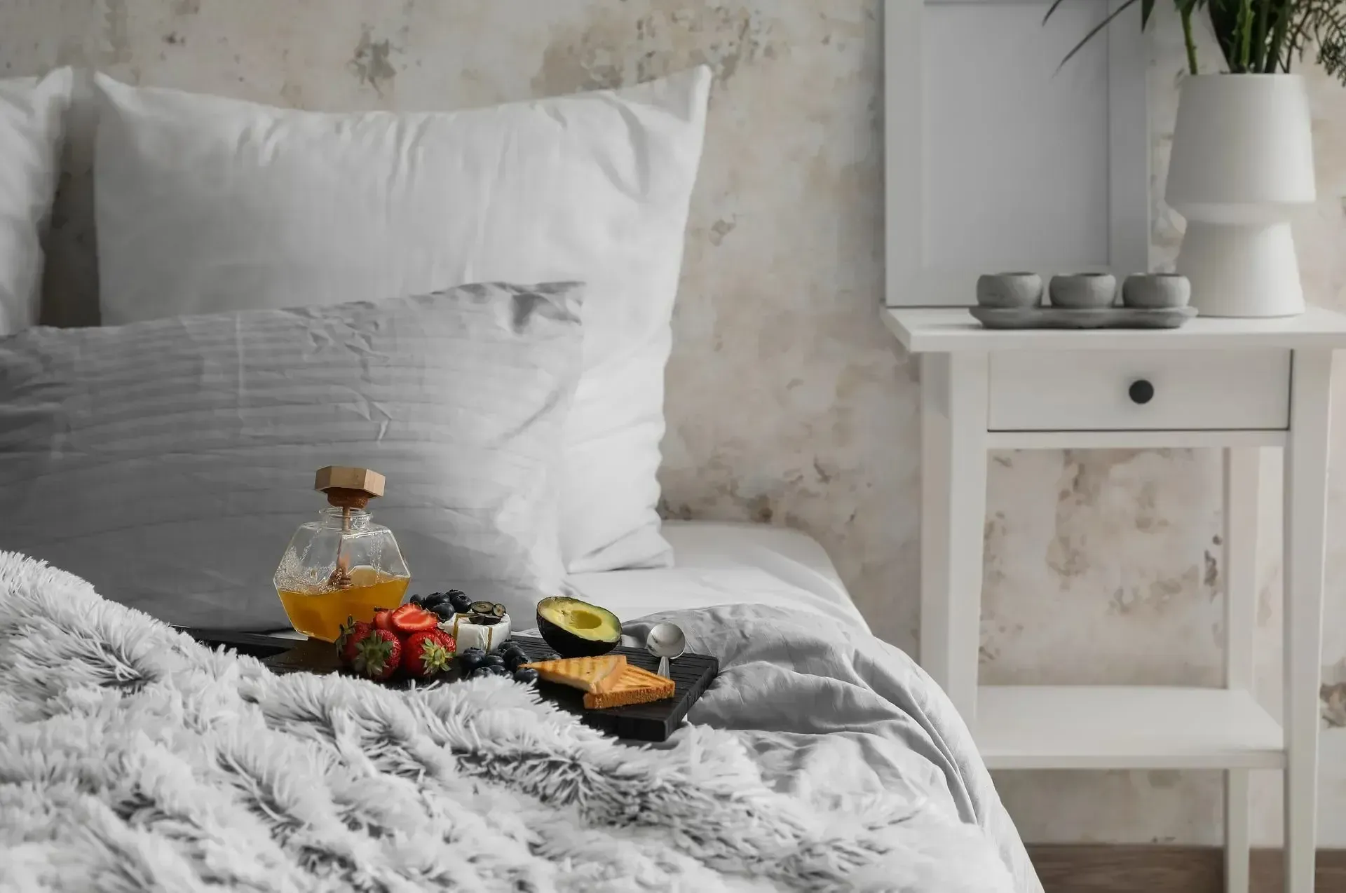 Breakfast on a tray with avocado toast, berries, and a carafe of honey resting on a bed with white pillows and a throw.