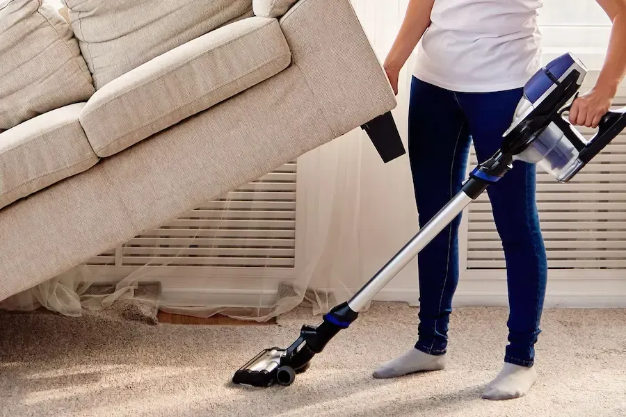 A person in a white shirt and blue jeans vacuums under a beige sofa using a cordless vacuum cleaner.