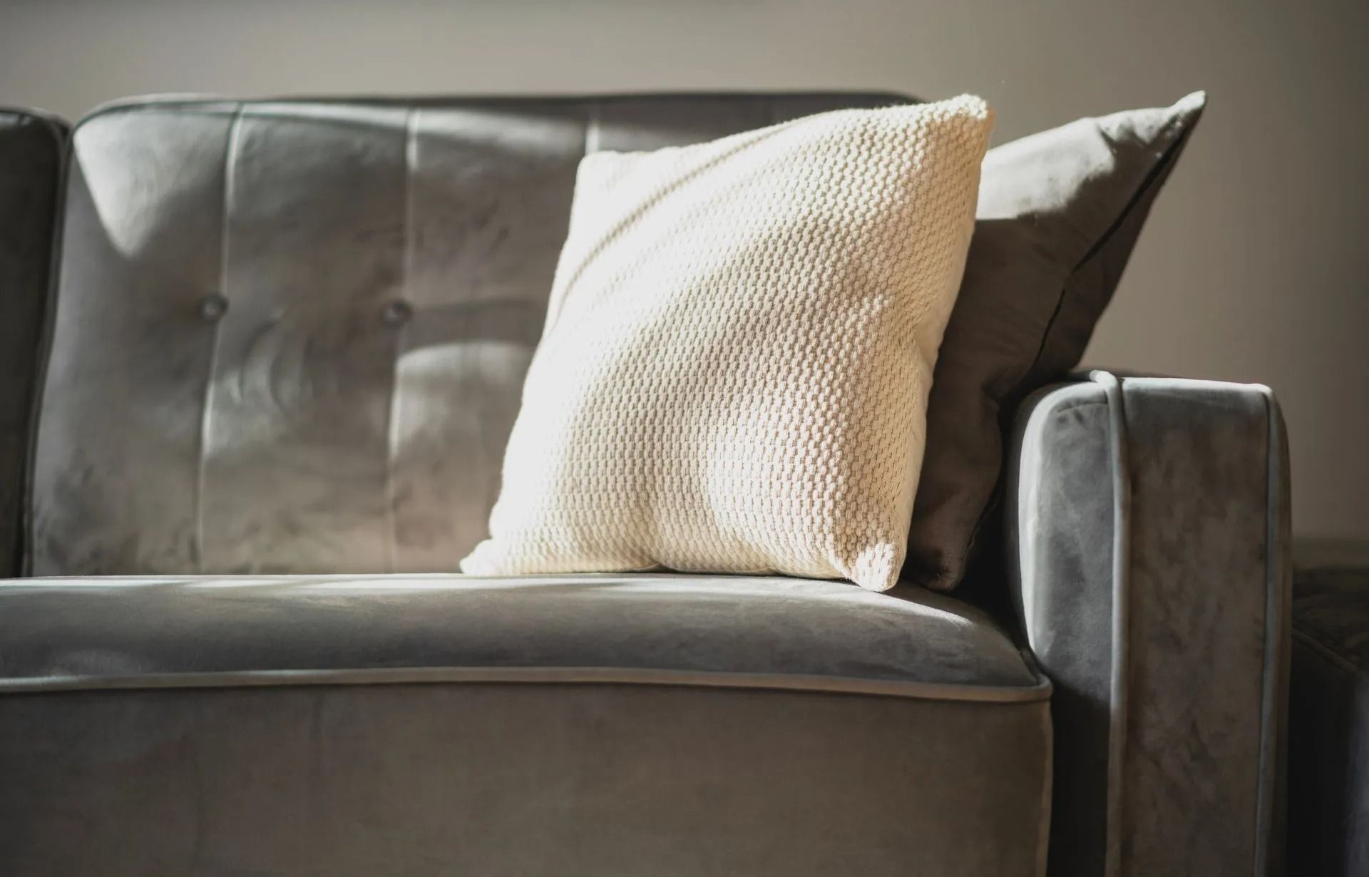 Gray velvet couch with a cream-colored knitted pillow in bright sunlight.