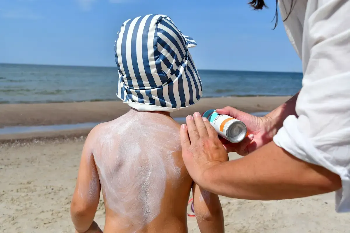 Person applying sunscreen to a child's back at the beach. Blue striped hat, blue ocean background.