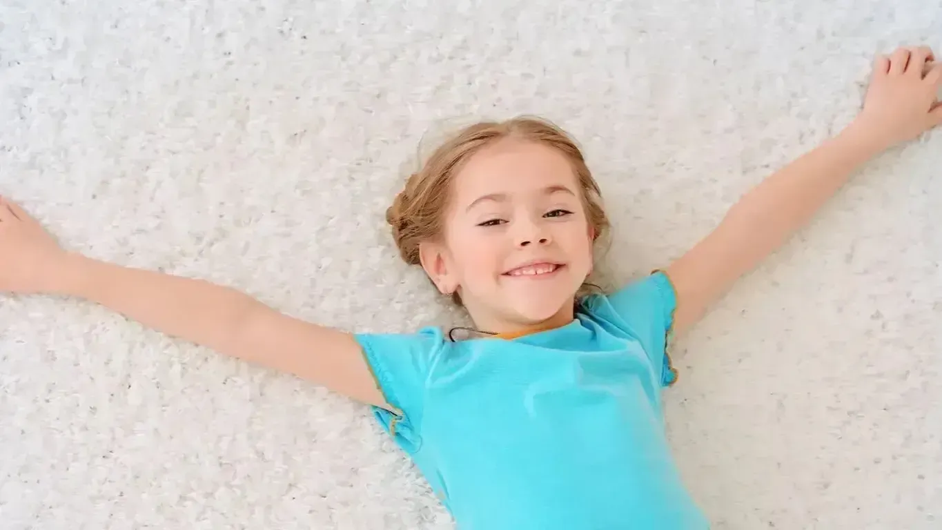 Girl with blonde hair, wearing a blue shirt, smiling with arms outstretched, lying on a white clean carpet.