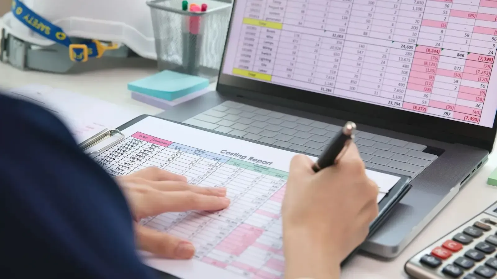 Person writing on a clipboard with a pen, looking at a spreadsheet on a laptop.
