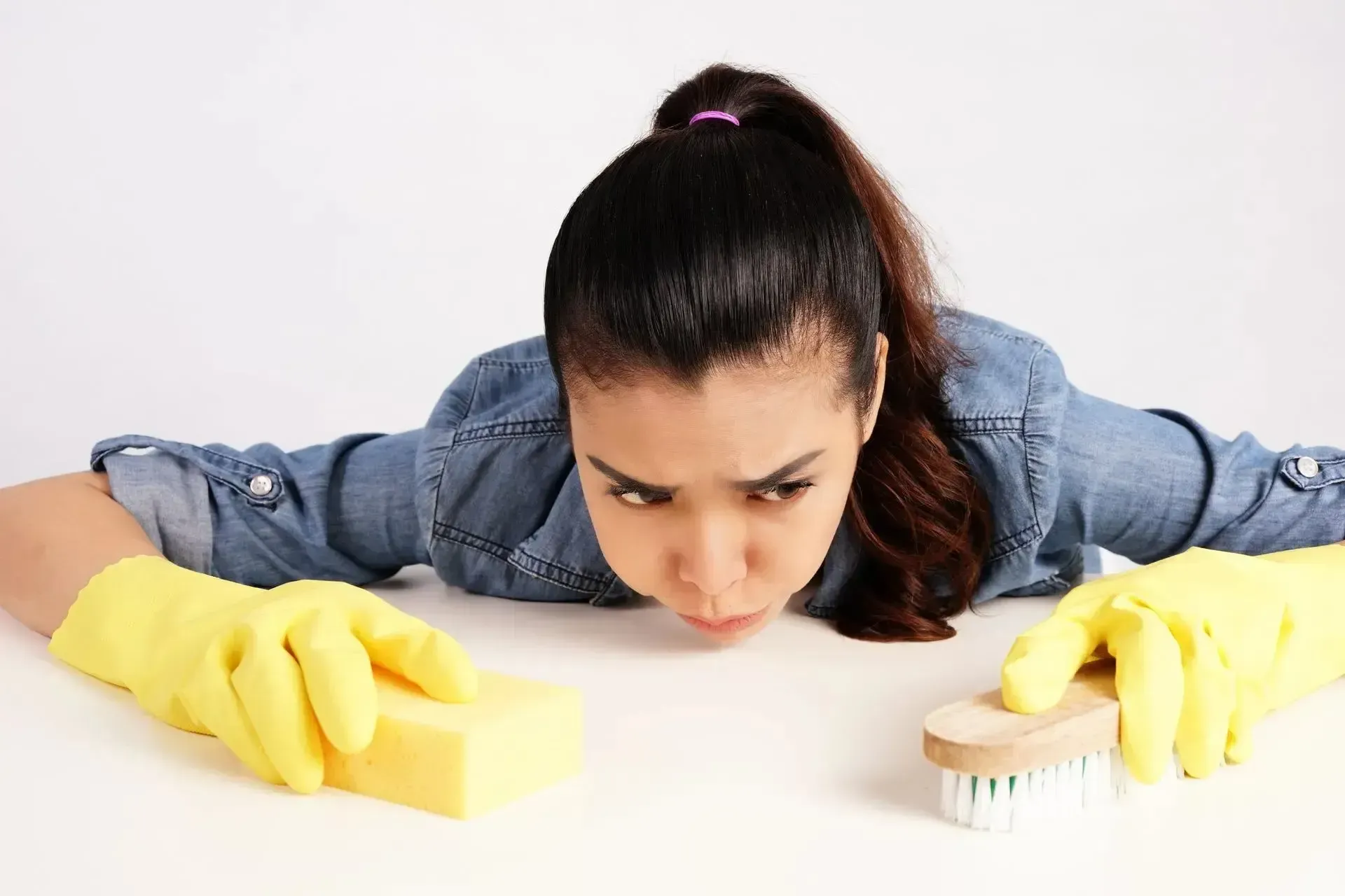 Woman wearing yellow gloves cleaning, leaning on a surface with a sponge and brush. She looks focused and frustrated.