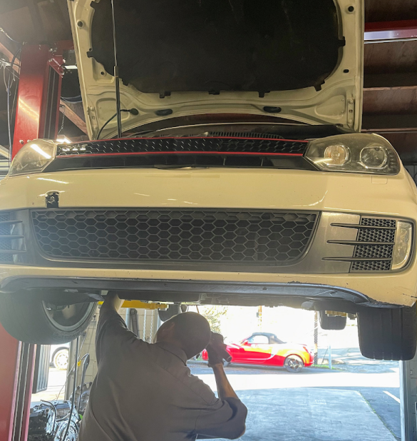 A mechanic works under a white car with an open hood on a lift. | Good Guys Auto Care & Tires