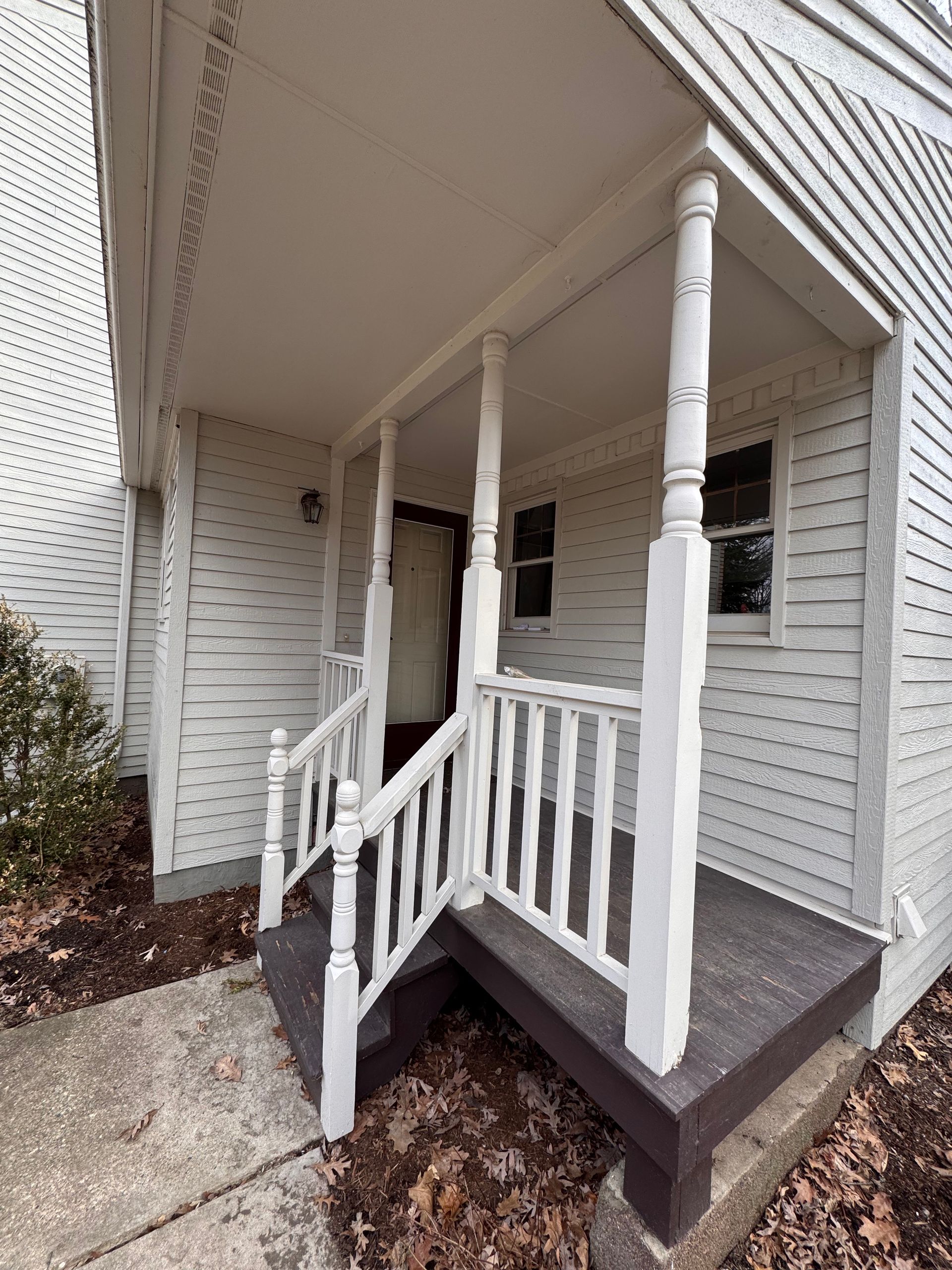 White porch with railing and columns, leading to a front door. Gray siding on the house.