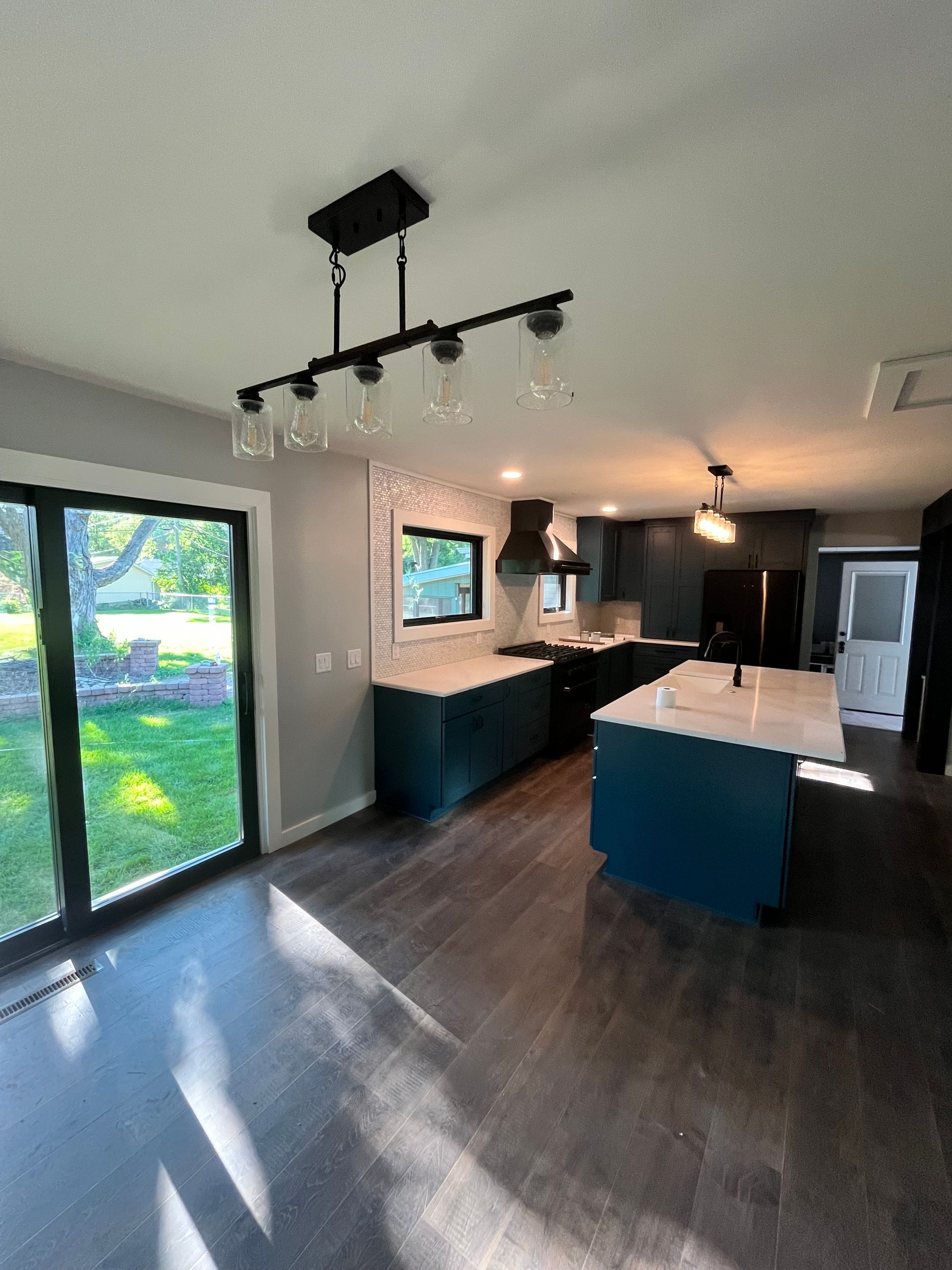 Modern kitchen with teal cabinets, white countertops, dark flooring, and a glass door leading to a yard.
