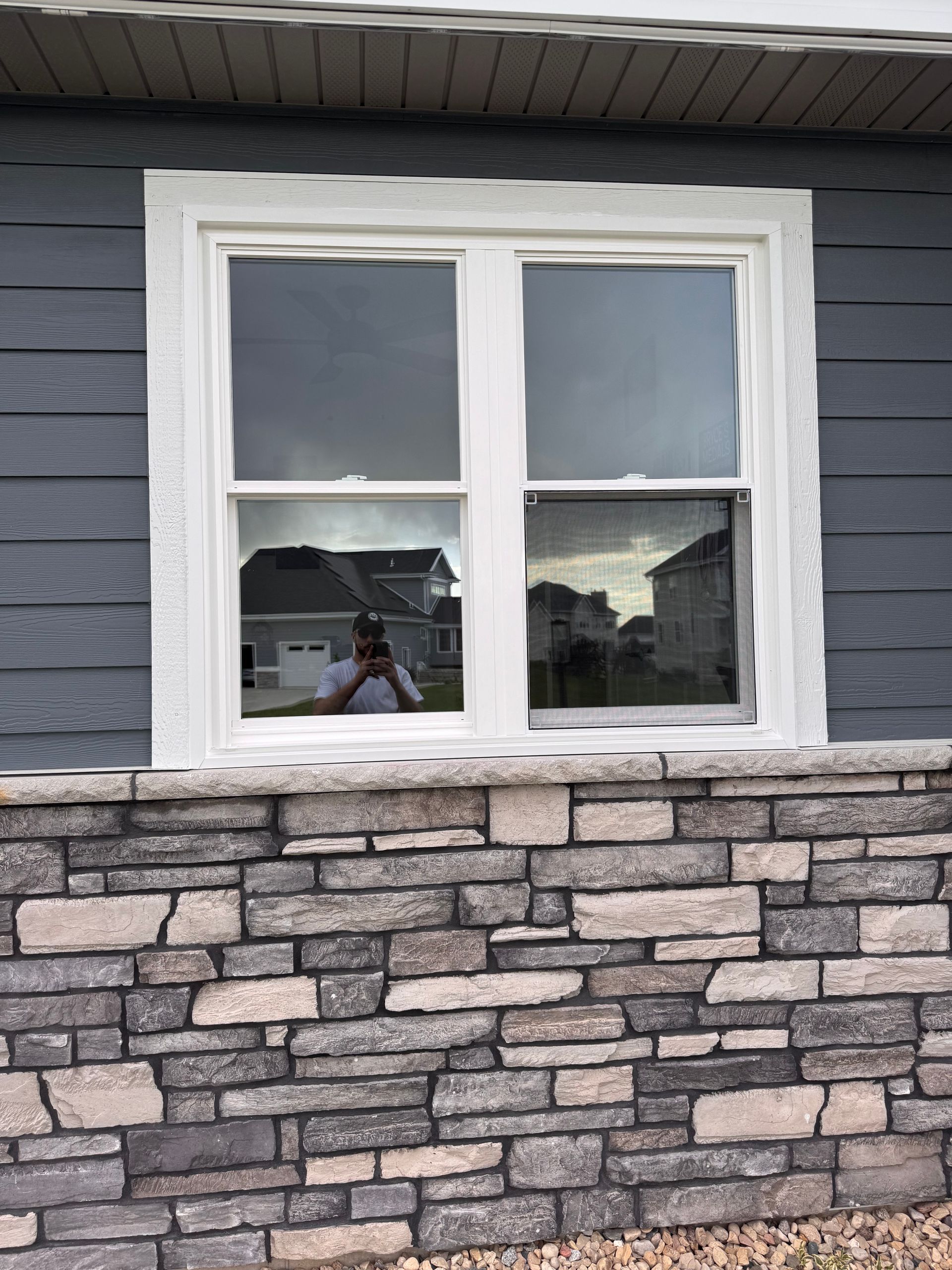 Window with white trim reflecting a house, sky, and person taking a photo; set in a gray and stone exterior wall.
