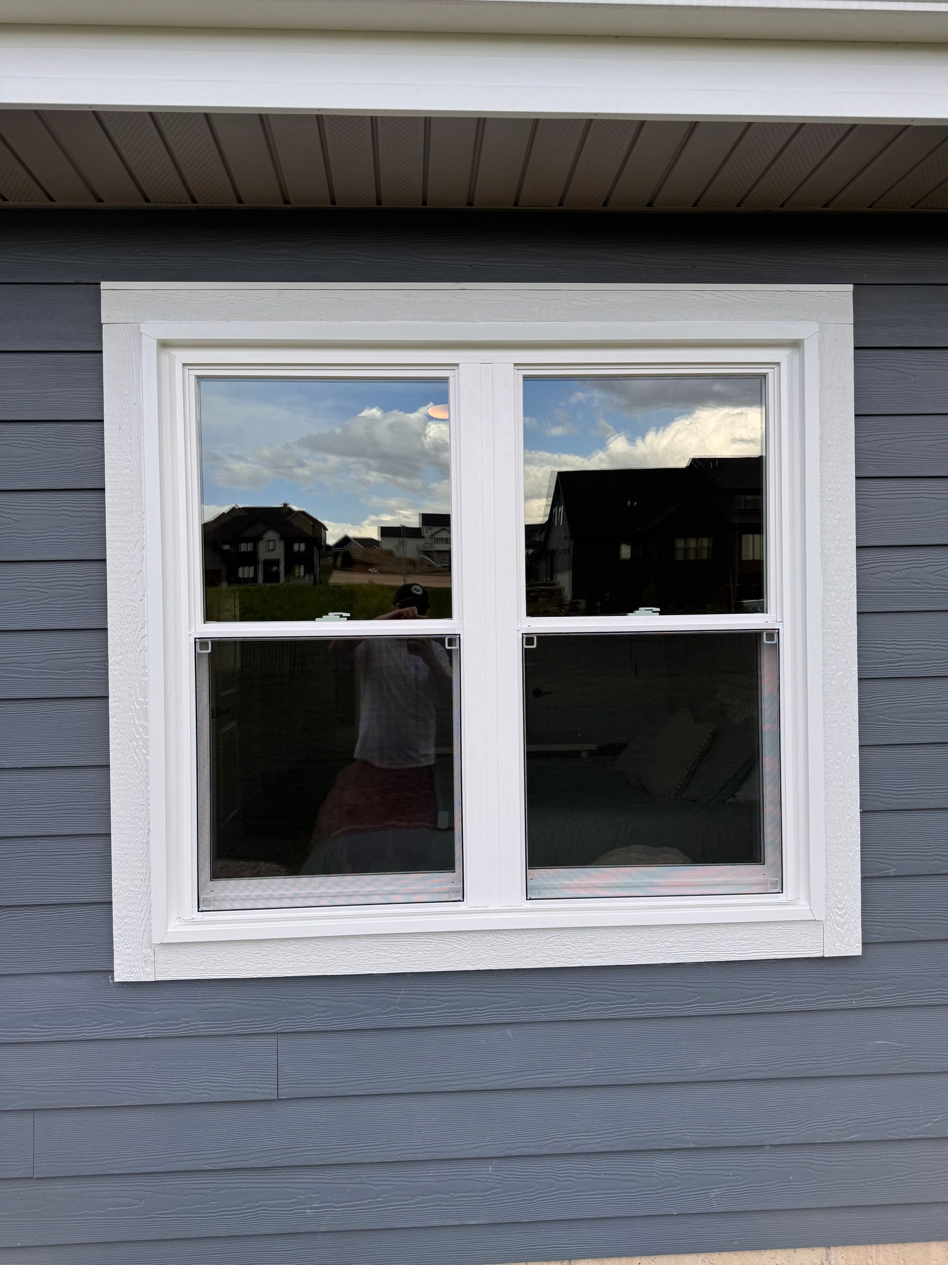 White-framed double-hung window on a gray building, reflecting blue sky and nearby houses.