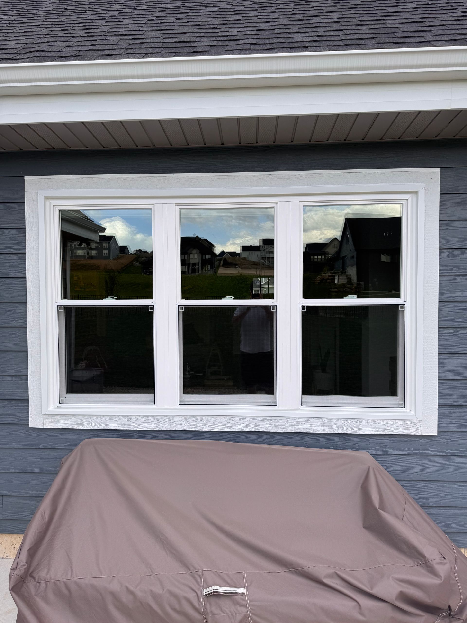 White-framed triple window set into a blue-gray house exterior, reflecting the outside, with a brown tarp below.