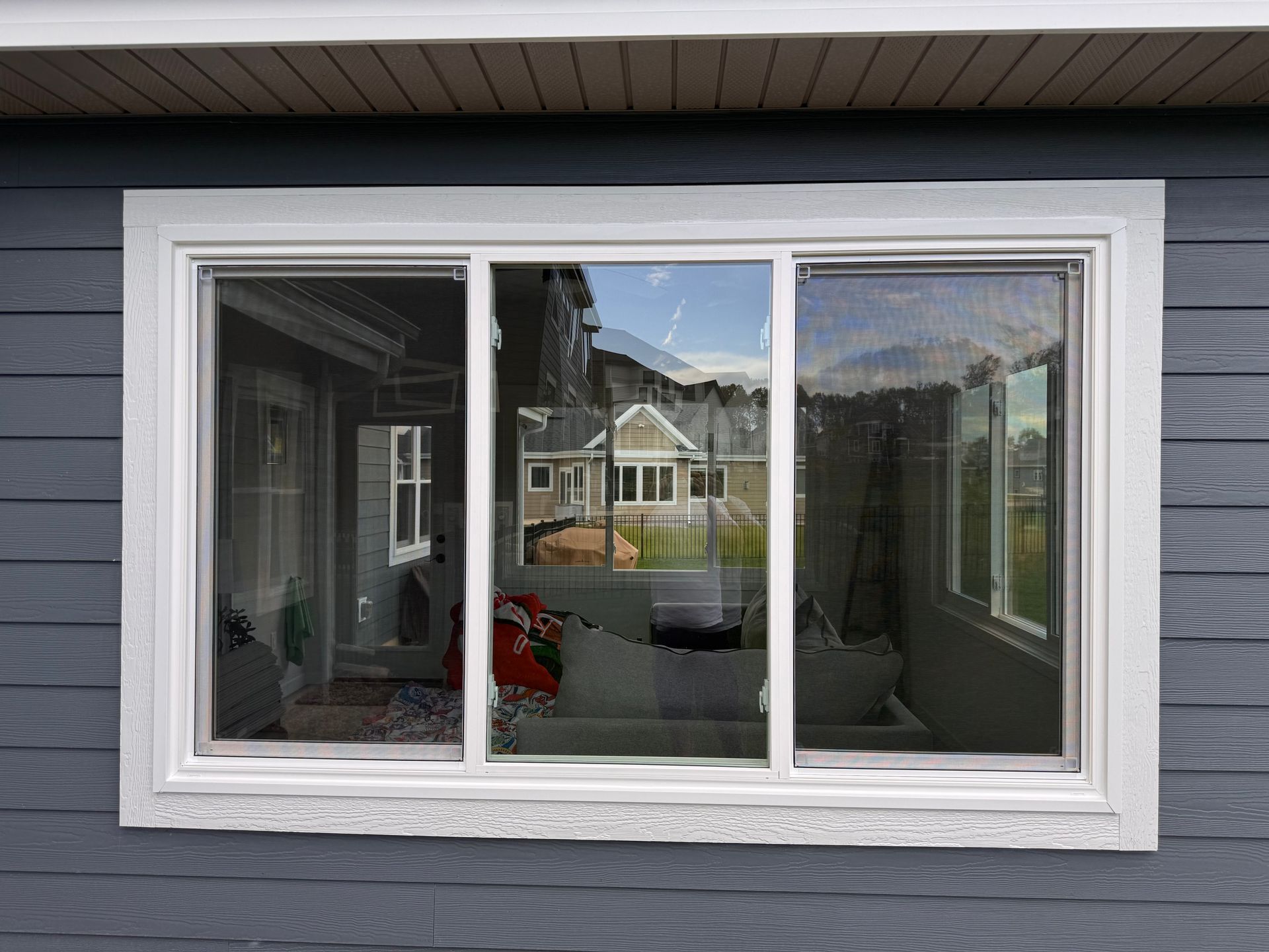 Window with white frame on blue siding reflects a house, furniture, and greenery.