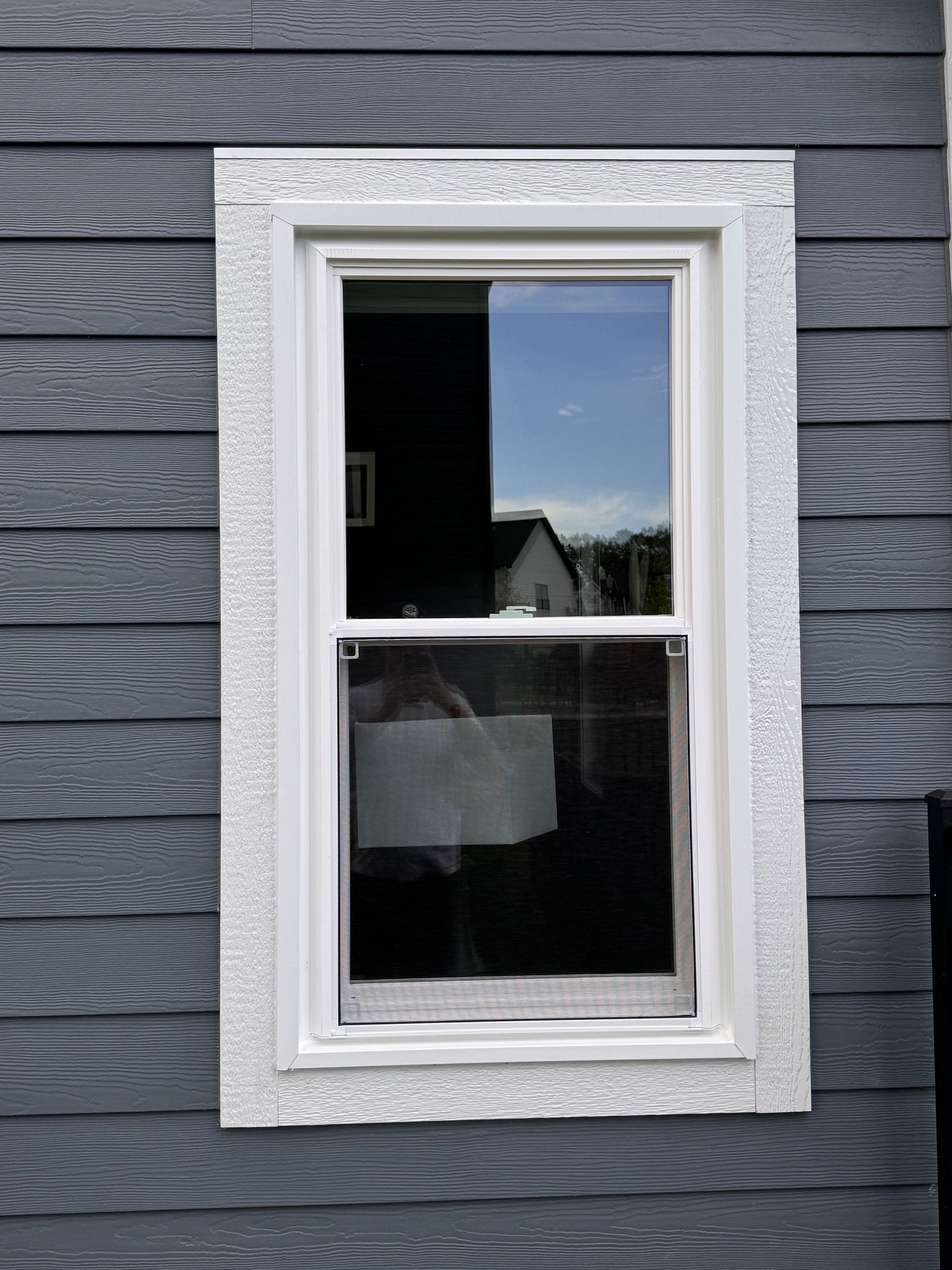 Window in a gray-sided building with white trim. The window is partially open, and reflects the sky.