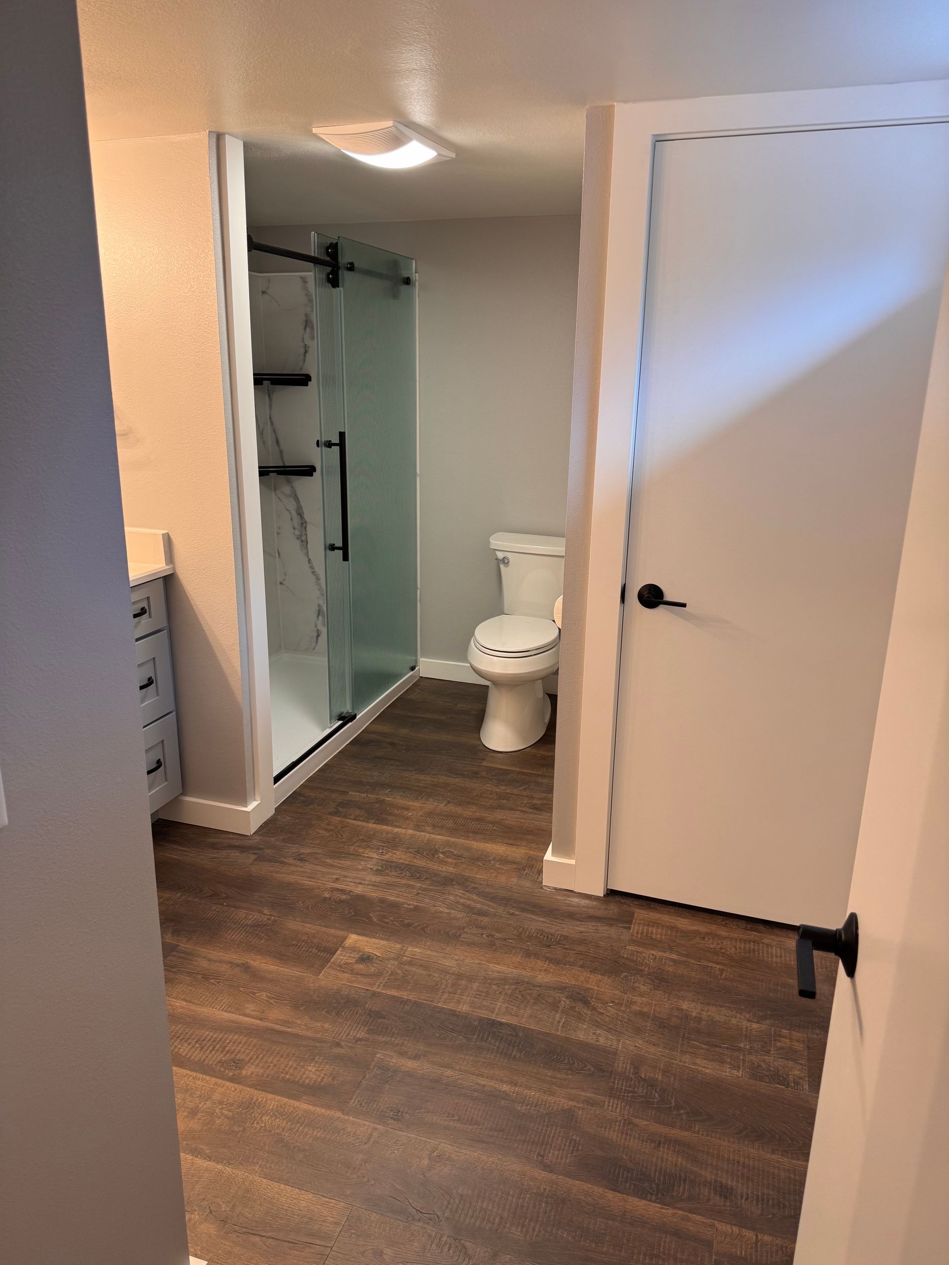 Bathroom with dark wood-look flooring, toilet, shower with black frame, and white door with black handle.