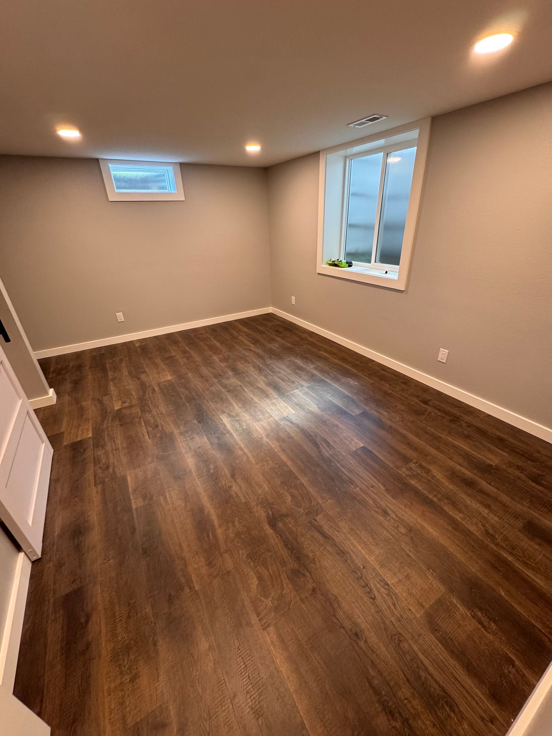 Empty room with dark wood-look flooring, neutral walls, two small windows, and recessed lighting.