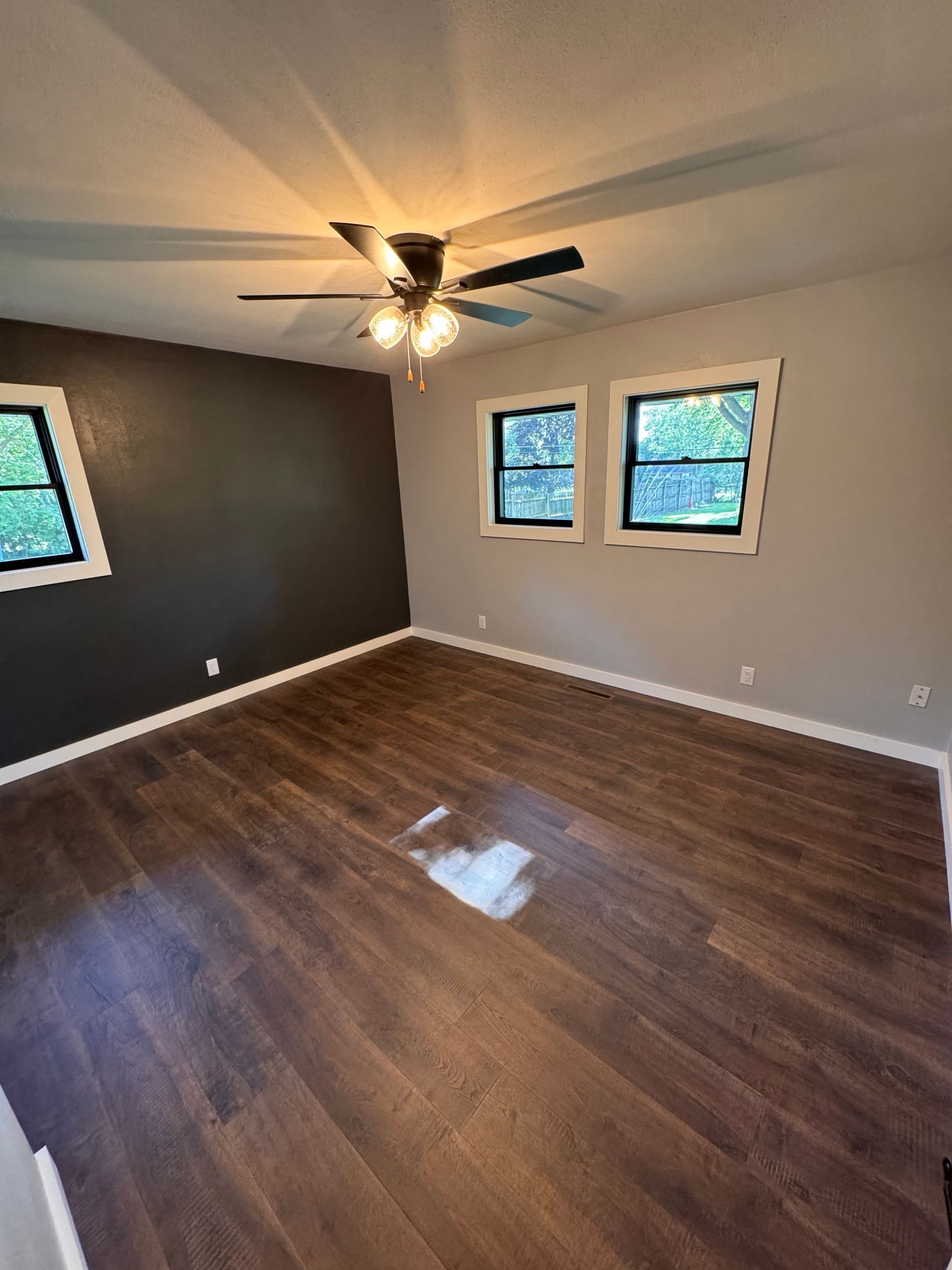 Empty bedroom with dark wood-look floor, gray walls, and black trim windows. A ceiling fan hangs above.
