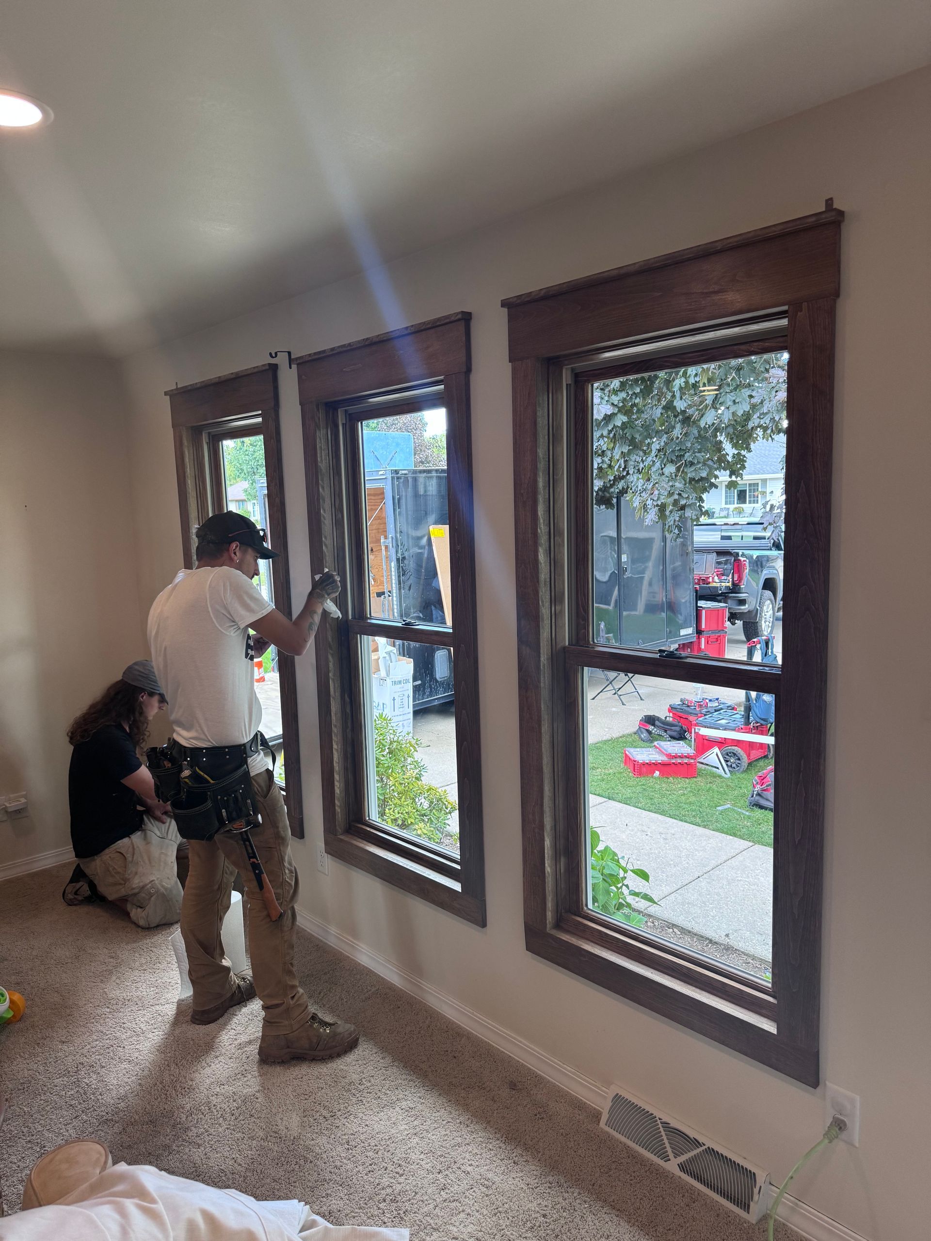 Two people installing window coverings on three windows. Interior shot with brown frames, beige walls and carpet.