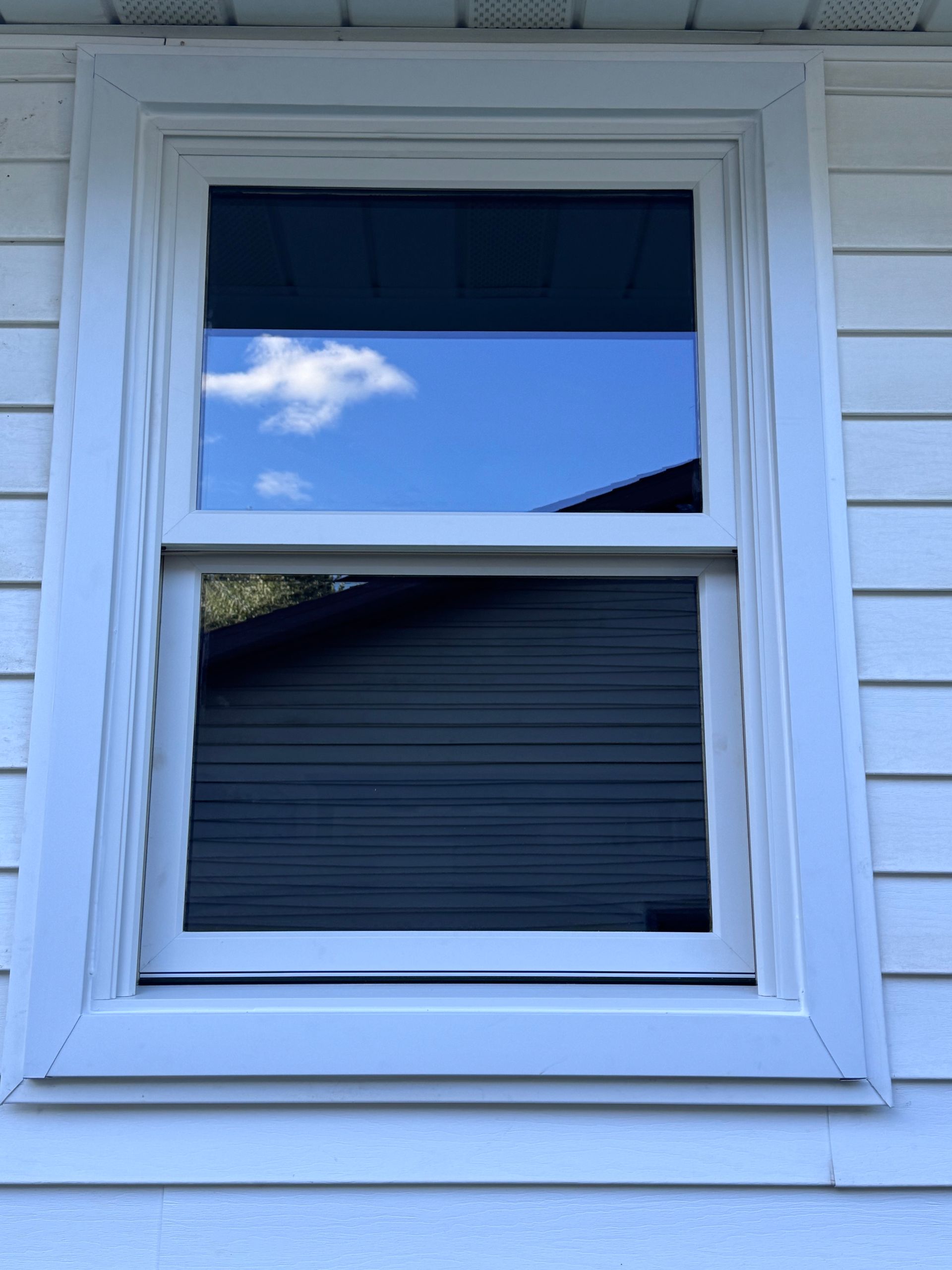White-framed window on a white siding exterior. The sky and a building are reflected in the glass.