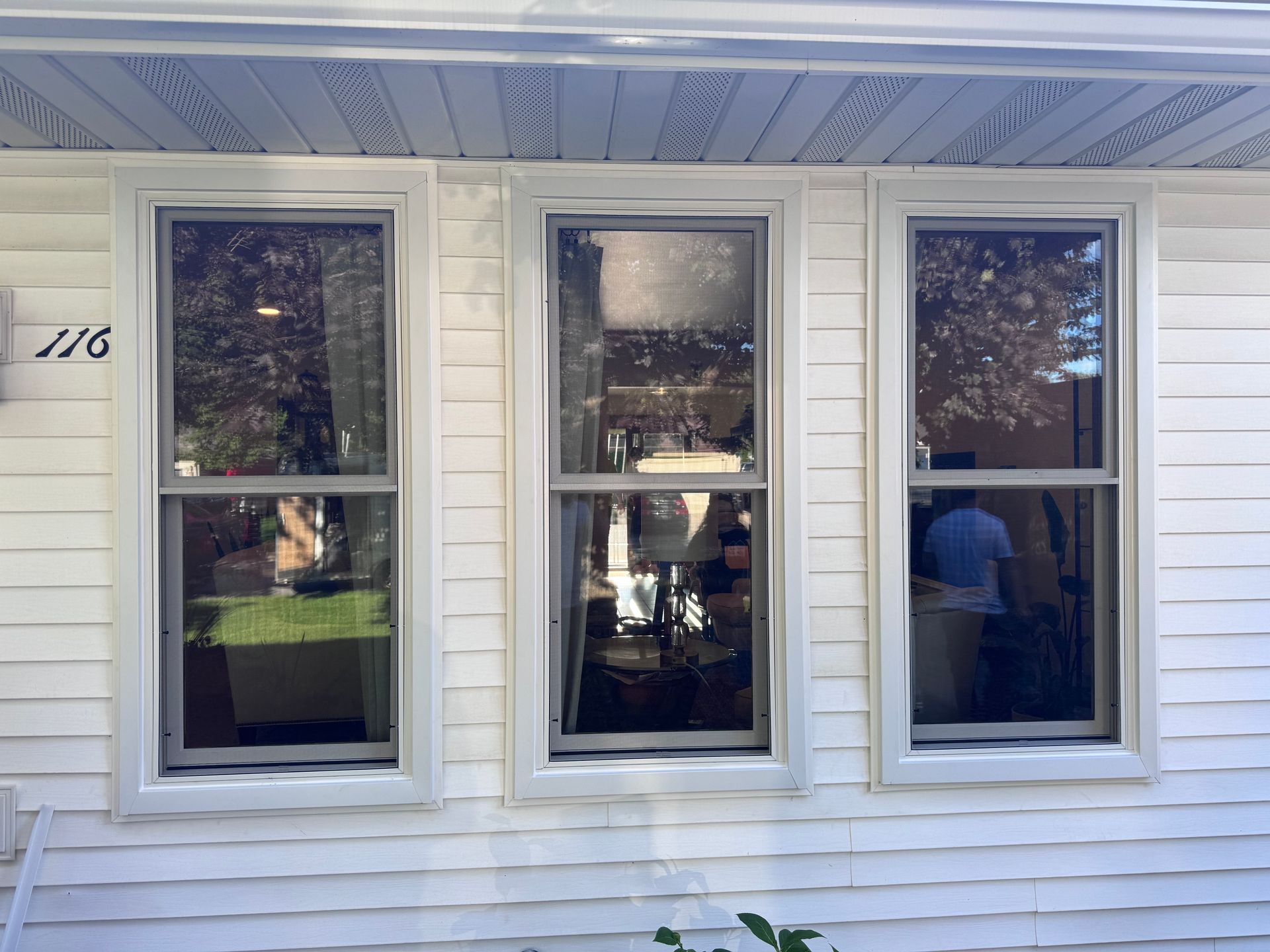 Three vertical windows with white frames on a white house exterior. The windows reflect outdoor scenes.