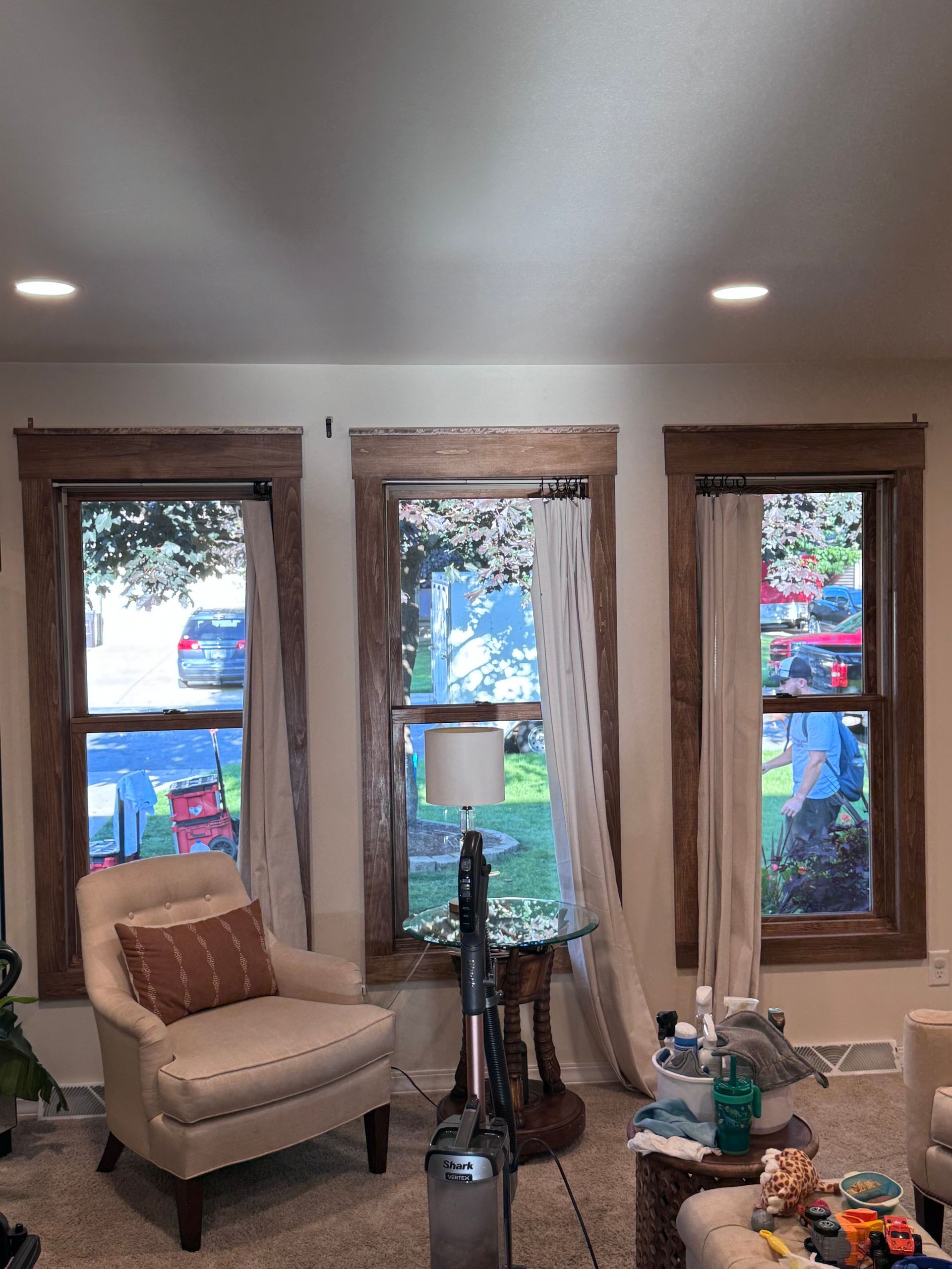 Three windows with brown trim, cream curtains, and a view of the outside. A beige chair and vacuum sit below the windows.