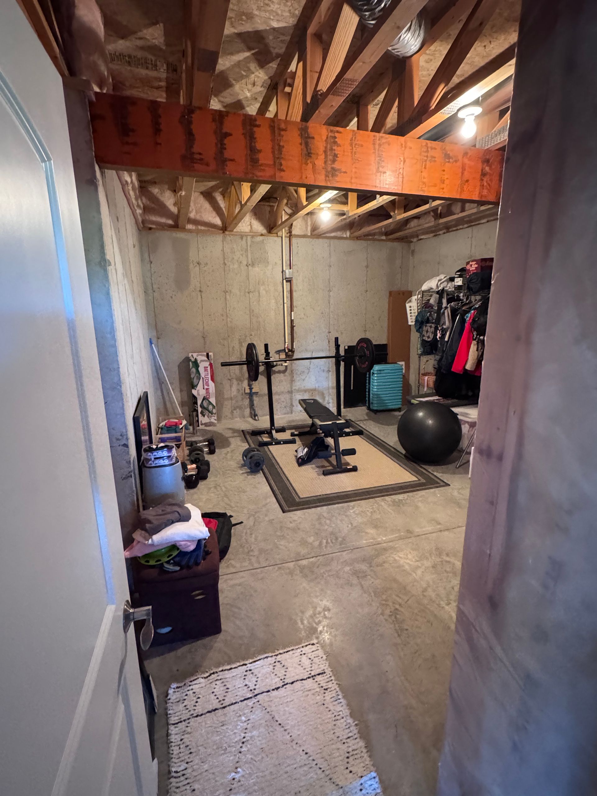Basement home gym with weight bench, barbell, and exercise equipment. Concrete walls, wood beams, and door frame.