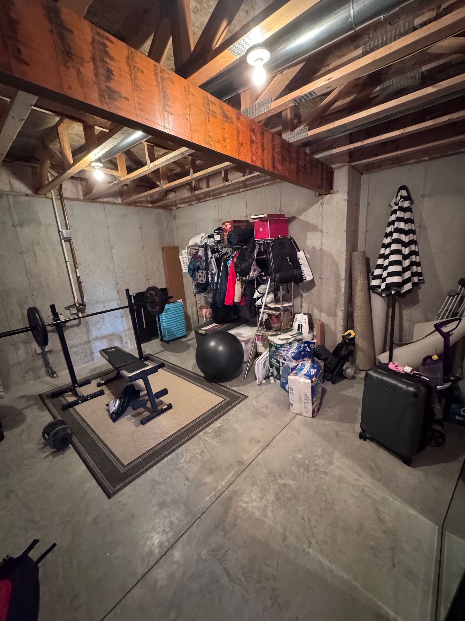 Basement with gym equipment, storage, and an umbrella, under exposed wooden beams and a light fixture.
