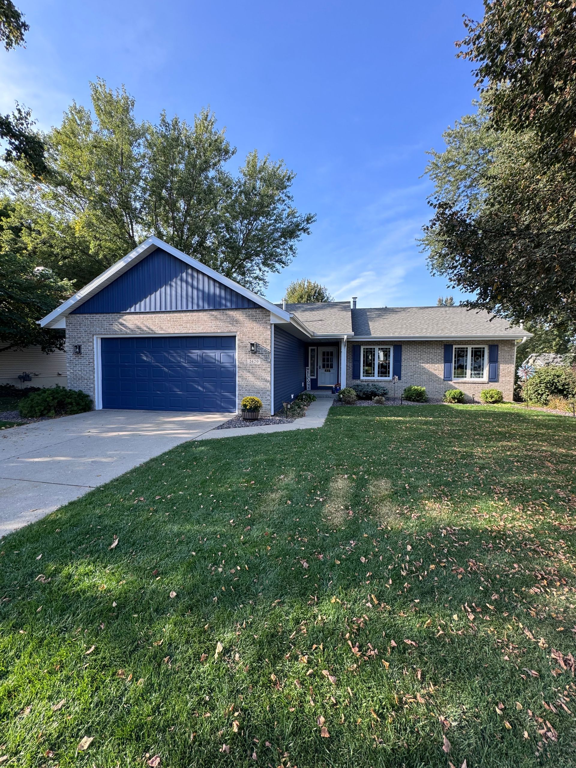 Blue-painted brick home with a blue garage door and trim, set on a green lawn with trees and a blue sky.