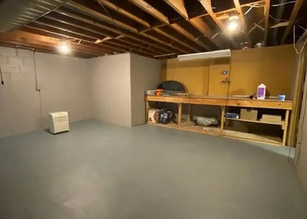Empty basement with gray painted walls and floor. Workbench and exposed ceiling.