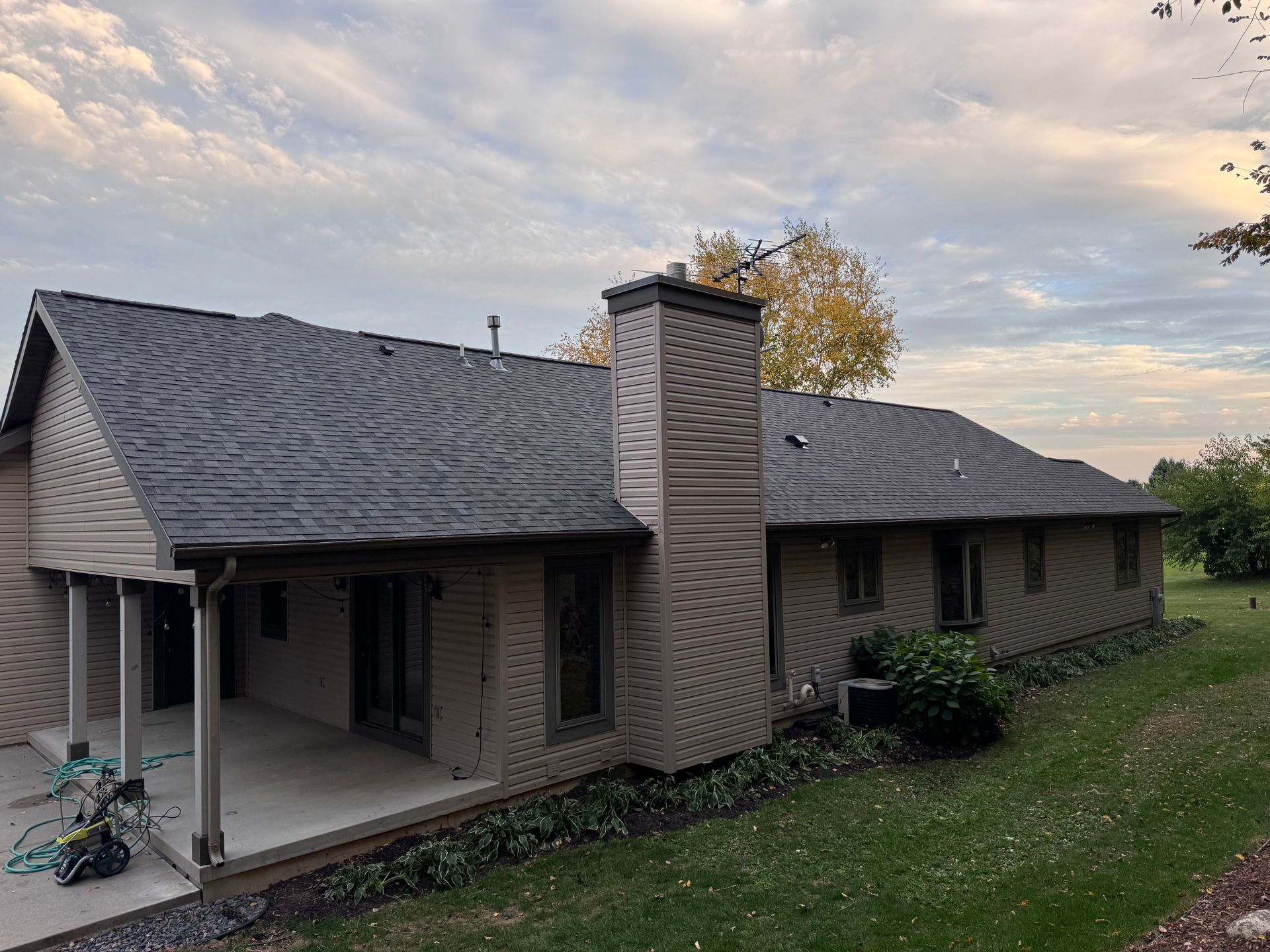 Tan house with dark gray roof, brick chimney, covered patio, and grassy yard under a cloudy sky.