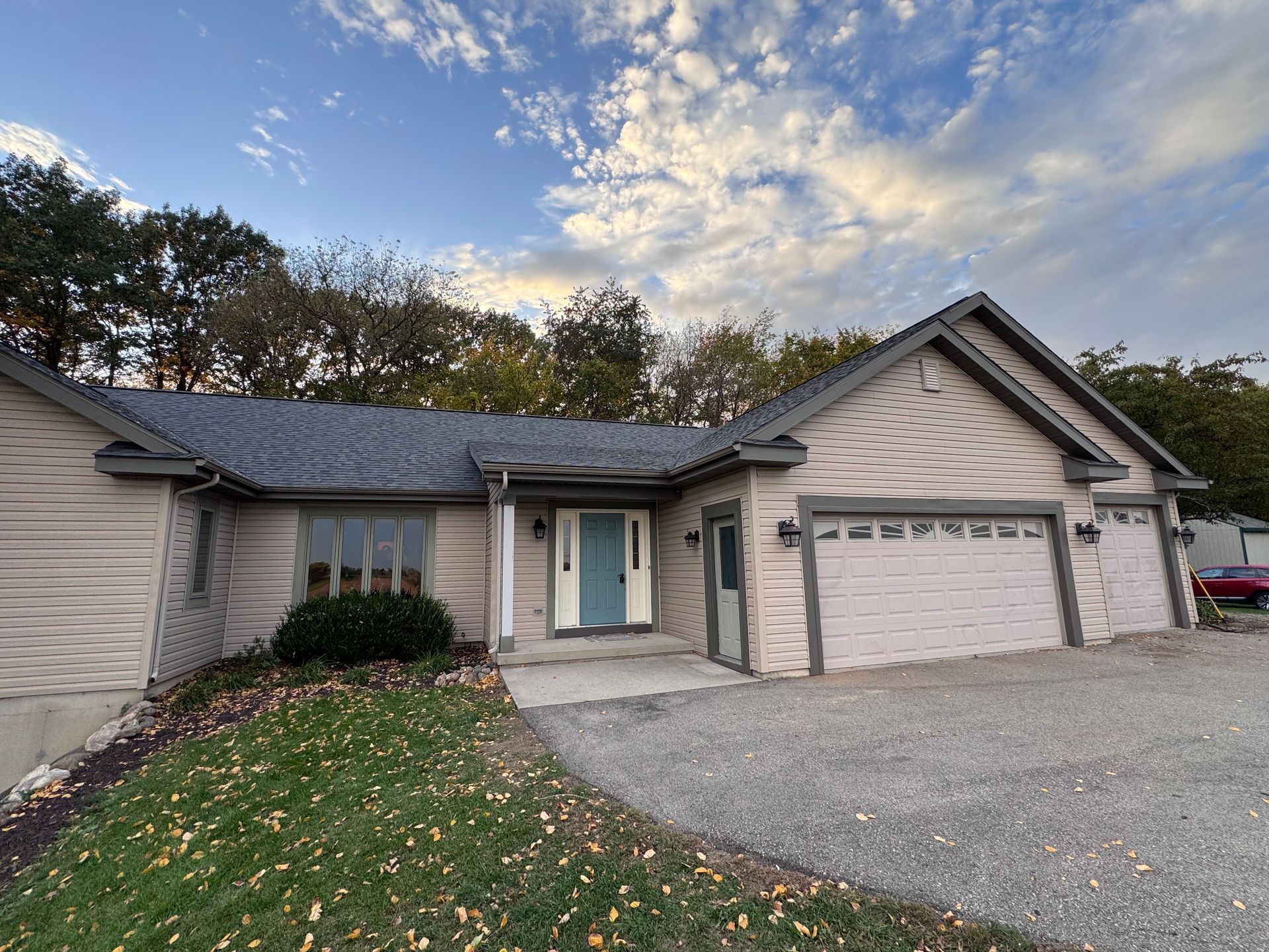 Beige house with a light blue door and two garage doors; gravel driveway, trees in the background, blue sky with clouds.