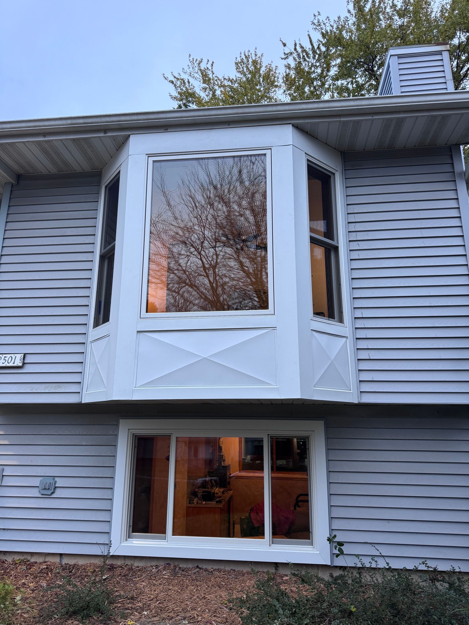 Two-story house with a white bay window and a rectangular window below, reflecting outdoor trees.
