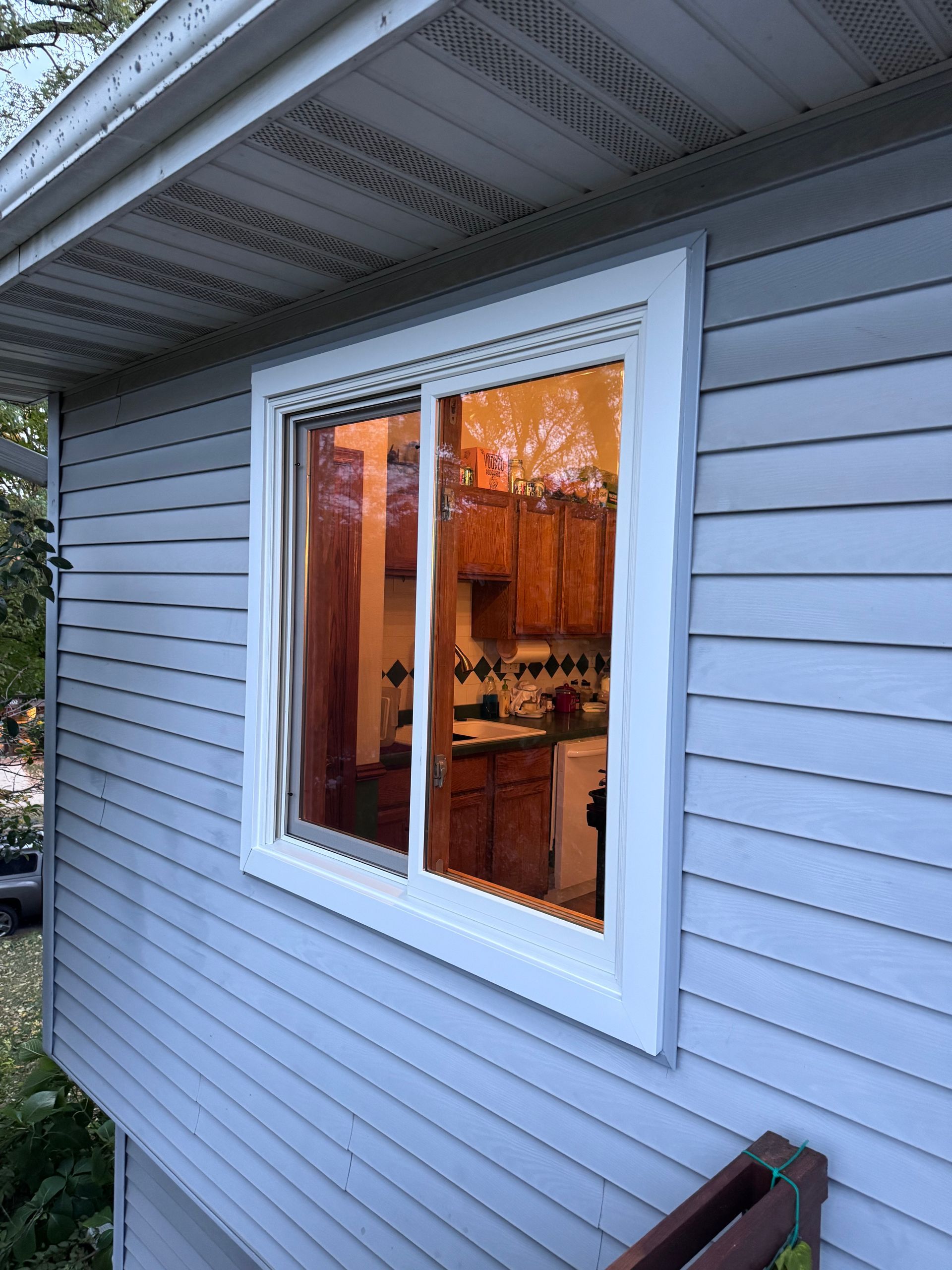 White-framed sliding window on a light blue-sided house, reflecting a kitchen with brown cabinets.