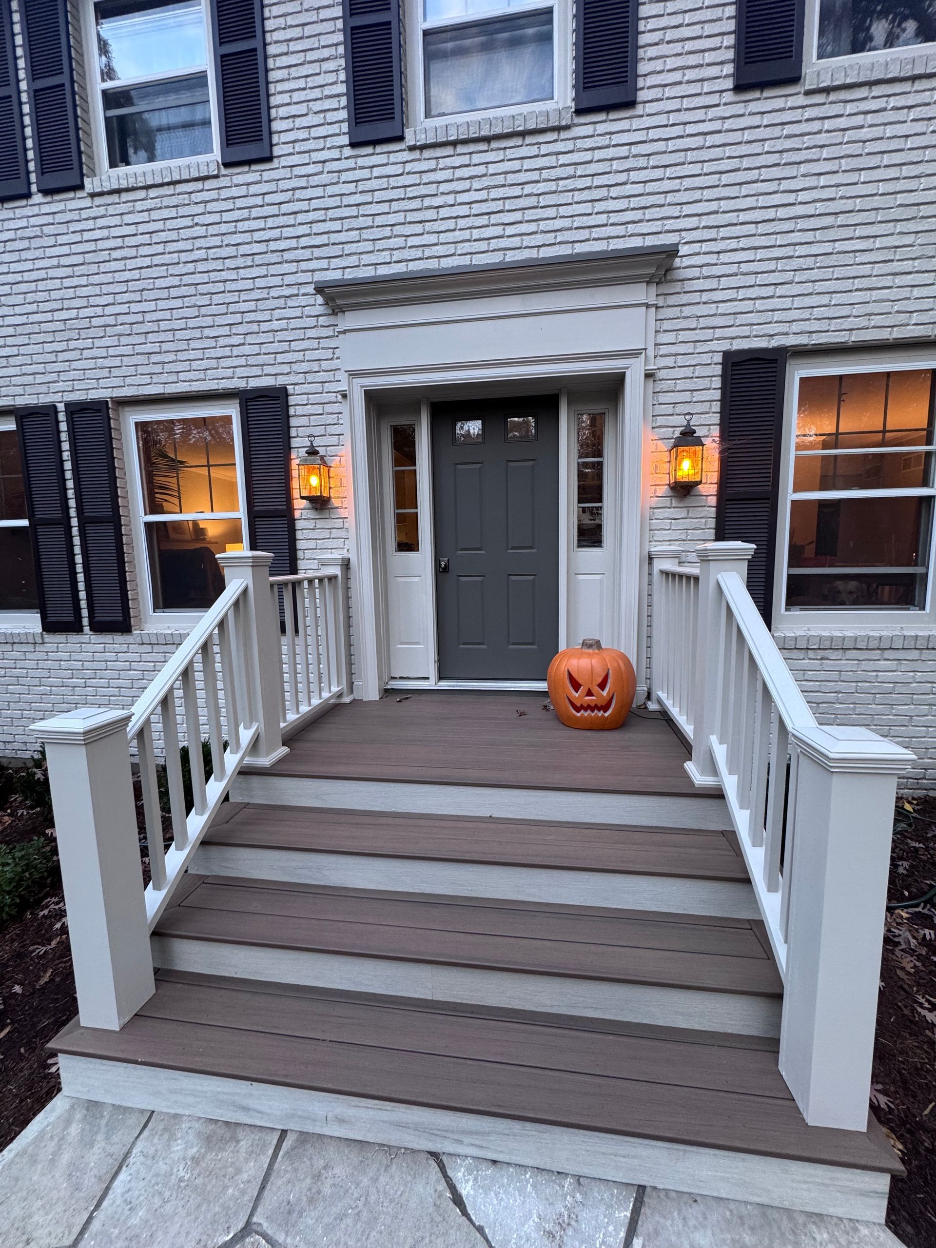 Brick house entrance with steps, pumpkin, and sconces. Door is gray, shutters are black.
