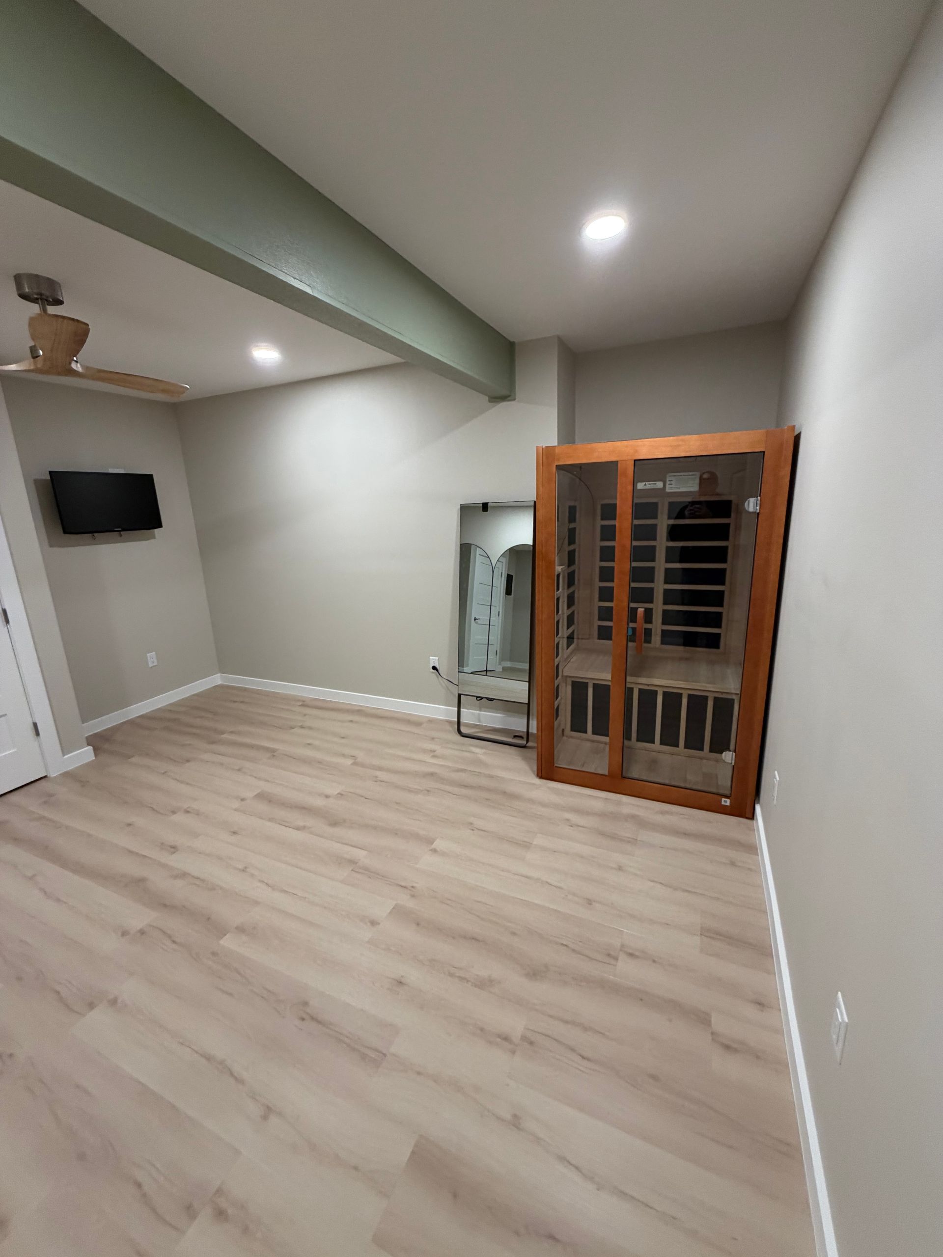Empty room with light wood flooring, a mirror, and a wooden sauna against a light gray wall.