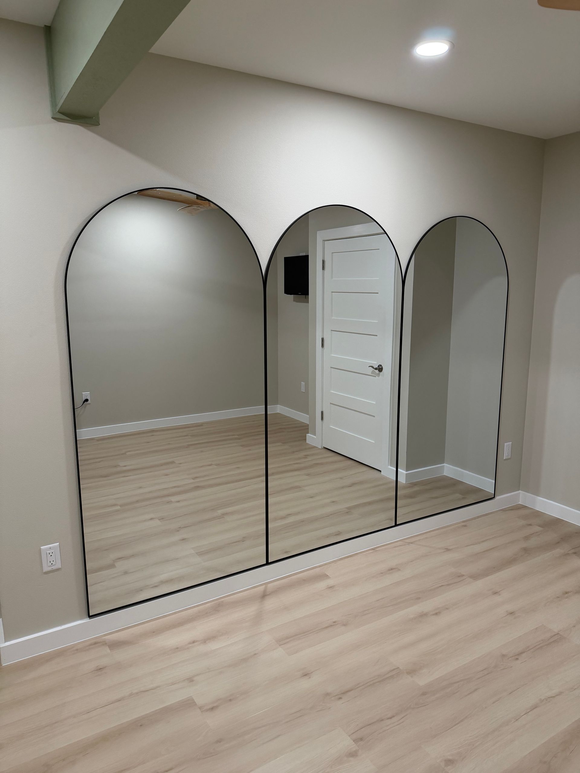 Three arched mirrors on a wall, reflecting a room with a door and wooden floor.