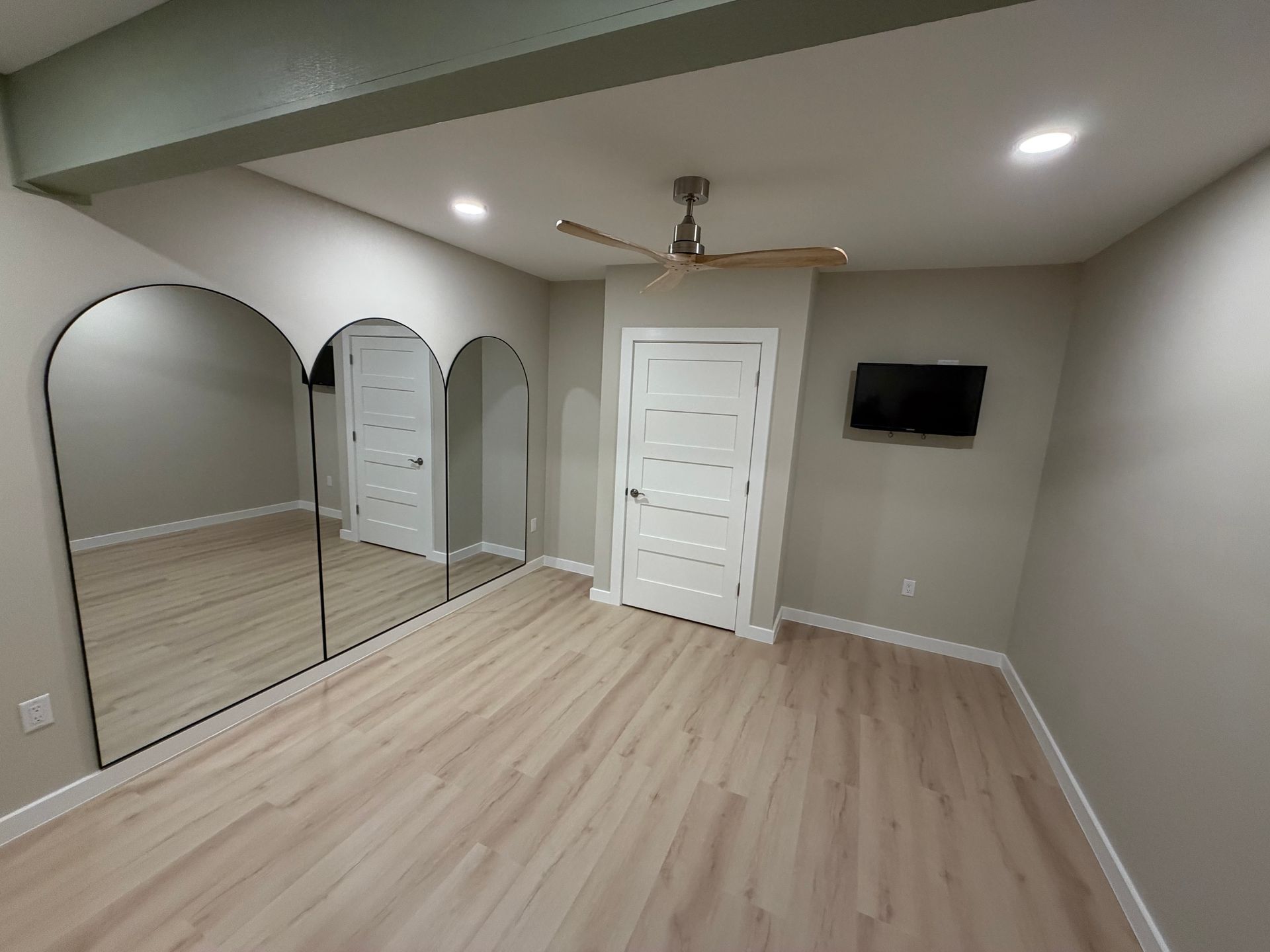 Empty bedroom with wood-look floors, large arched mirrors, white door, TV, and ceiling fan. Walls are light gray.