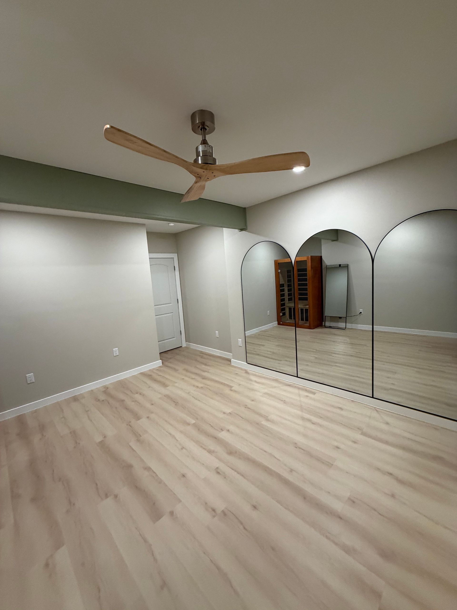 Empty room with light wood floors, mirrors, and a ceiling fan. Walls are gray and a soft green.