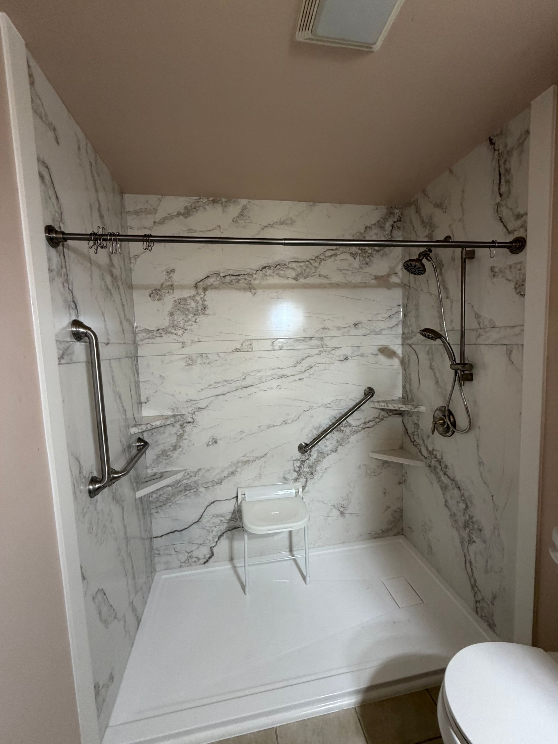 Accessible shower with marble-look walls, grab bars, shower seat, and handheld showerhead.