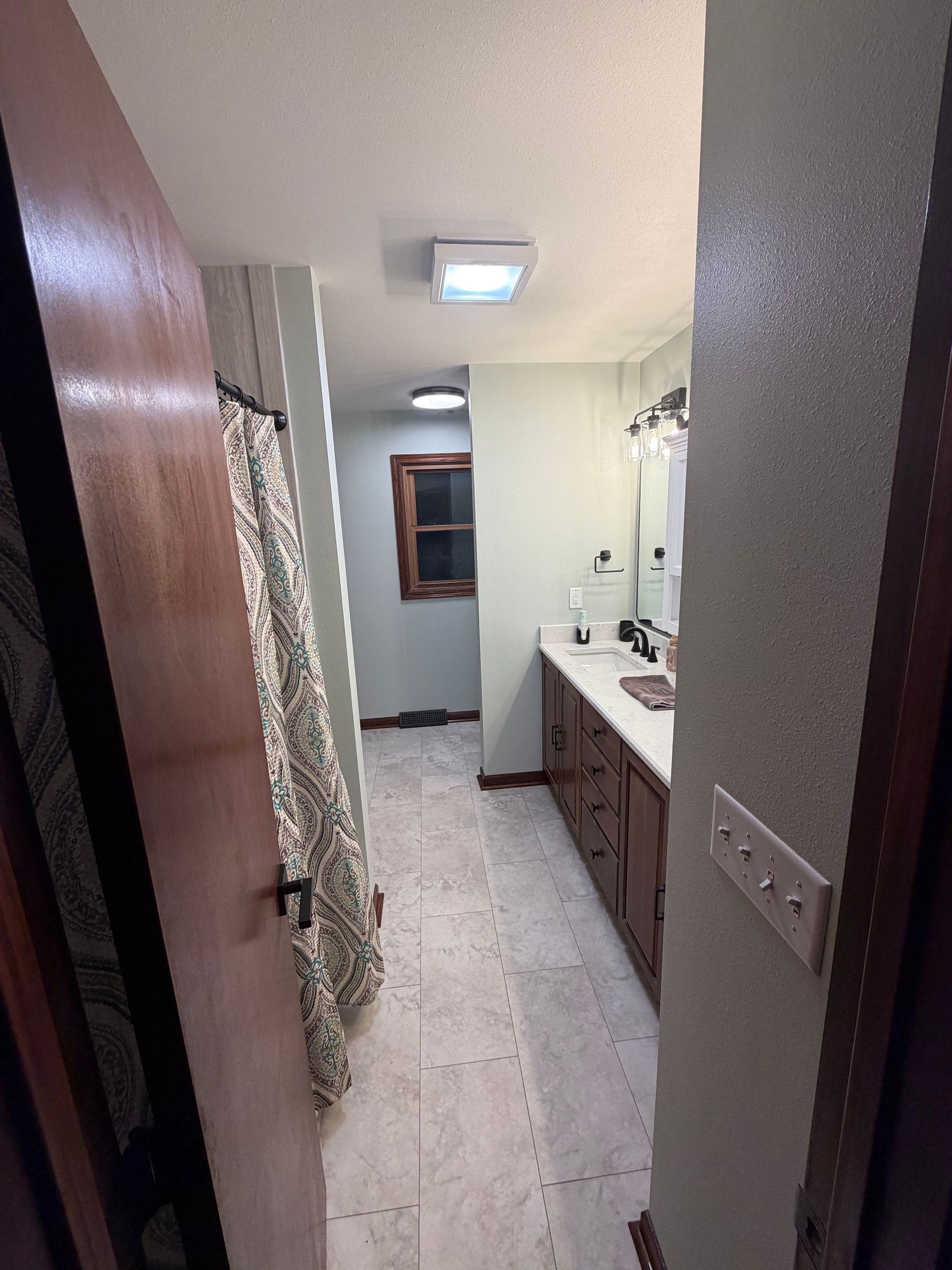 Narrow bathroom with open door. Light green walls, brown cabinets, and a window.