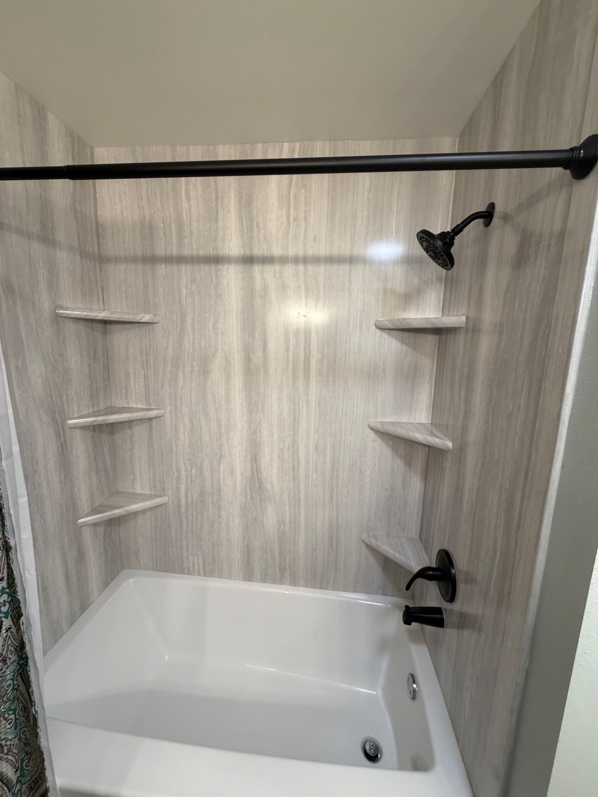 Bathroom with white bathtub, showerhead, shelves, and gray wall panels. Black shower rod and faucet.