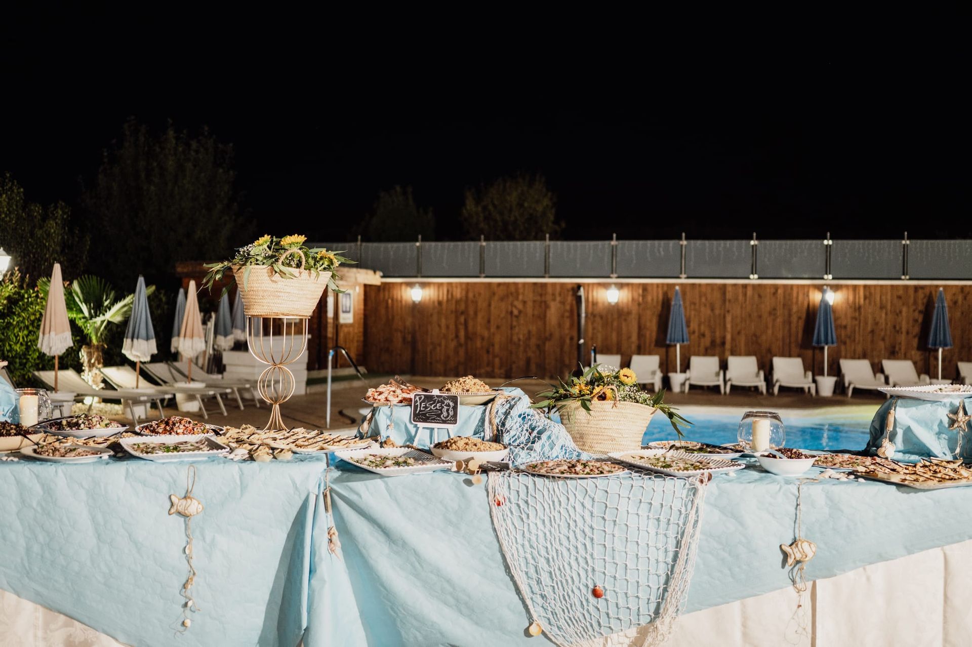 Tavolo da buffet serale allestito vicino alla piscina, decorato con frutti di mare e cestini floreali, con dettagli blu.