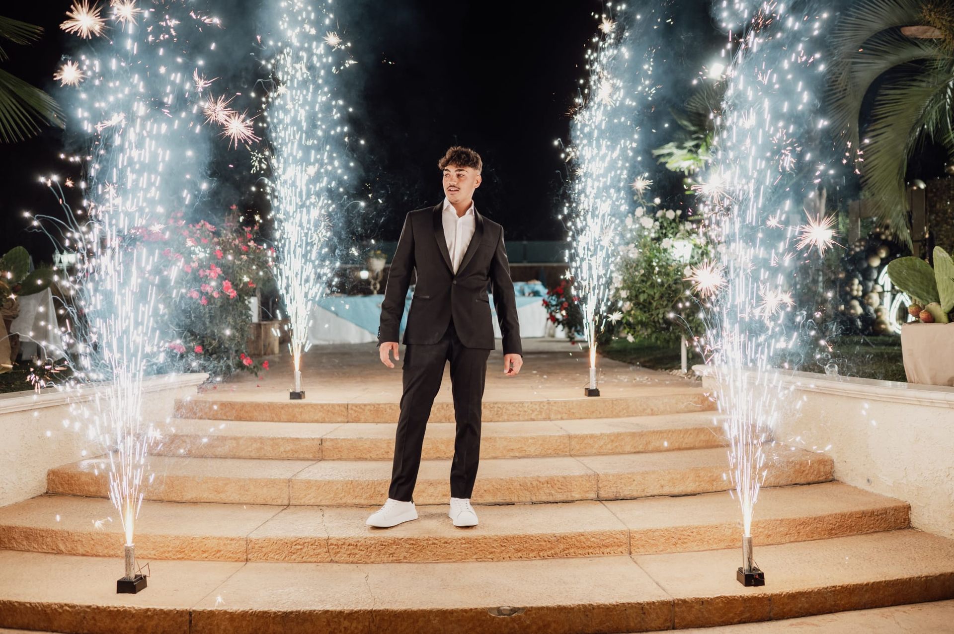 Un uomo in abito nero è in piedi sui gradini tra due fontane di scintille di notte.