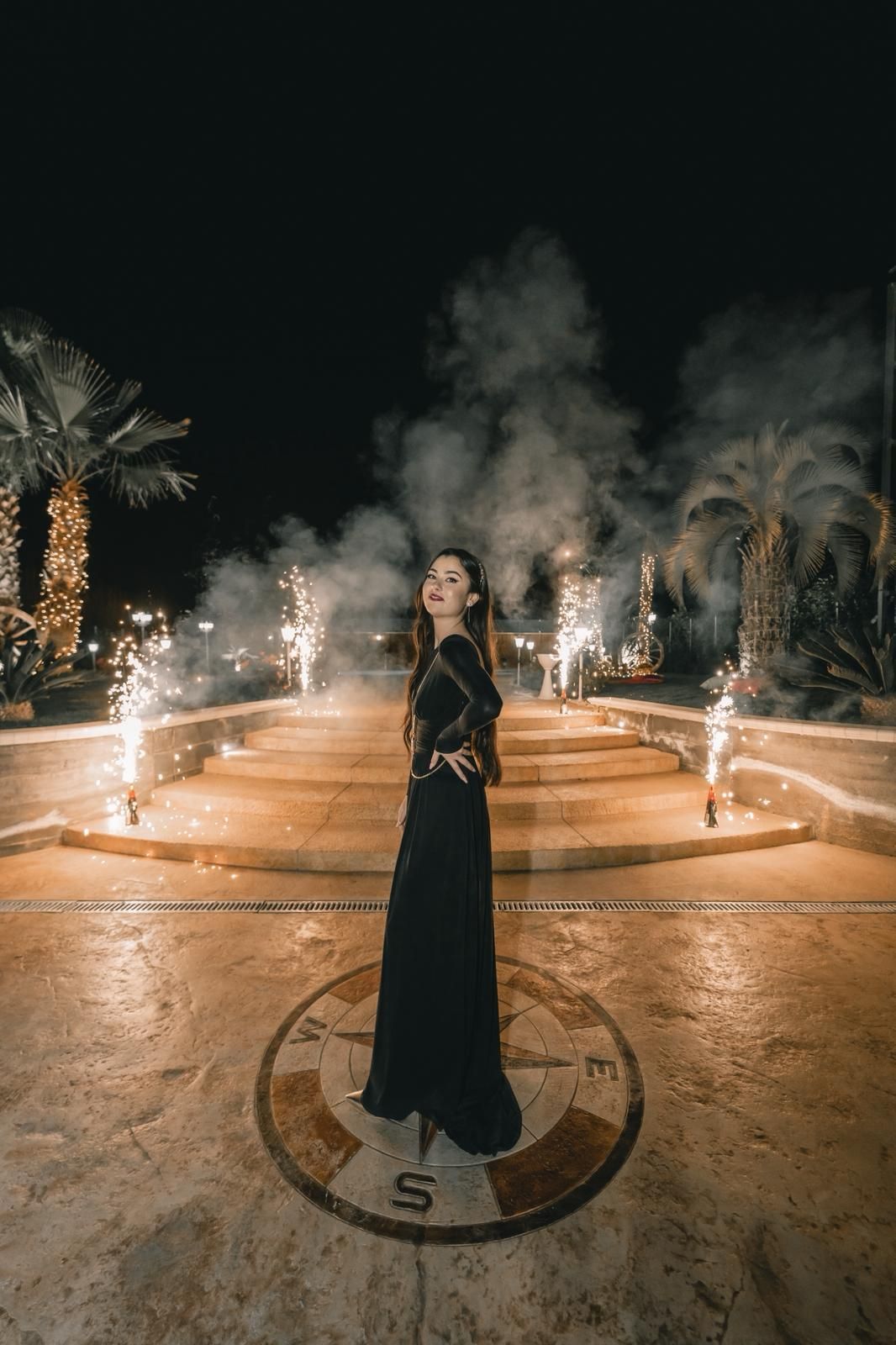 Una donna in abito nero posa di notte su una bussola, fuochi d'artificio e palme illuminate.