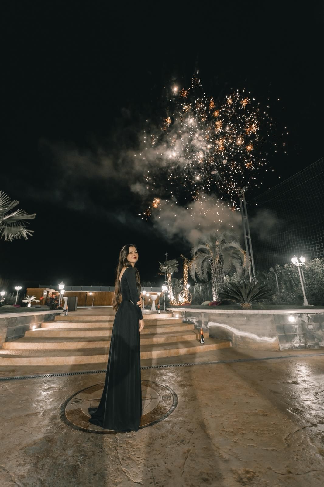 Una donna in abito nero guarda i fuochi d'artificio di notte. Scenario all'aperto con gradini, palme e luci.
